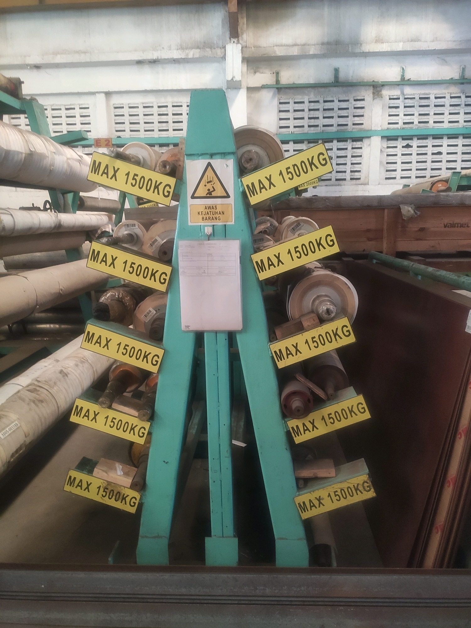 The image shows a storage area with industrial materials, likely in a warehouse or factory. The foreground is dominated by a teal-colored metal rack holding several large rolls of material, possibly paper or fabric. Yellow warning signs with "MAX 1500KG" are attached to the rack at various points, indicating weight limits. To the right of the rack, a stack of large, dark brown flat materials, possibly metal sheets or wood panels, is leaning against a wooden structure. In the background, a wall with white horizontal louvers is visible, and overhead fluorescent lighting fixtures are present. A warning sign with a black triangle and an exclamation mark, along with falling person symbol, is attached to the teal rack, with text below that reads "AWAS KEJATUHAN BARANG" which translates to "WARNING FALLING OBJECTS". The environment appears to be an indoor industrial setting, with a concrete floor visible at the bottom. There are no people present in the image.