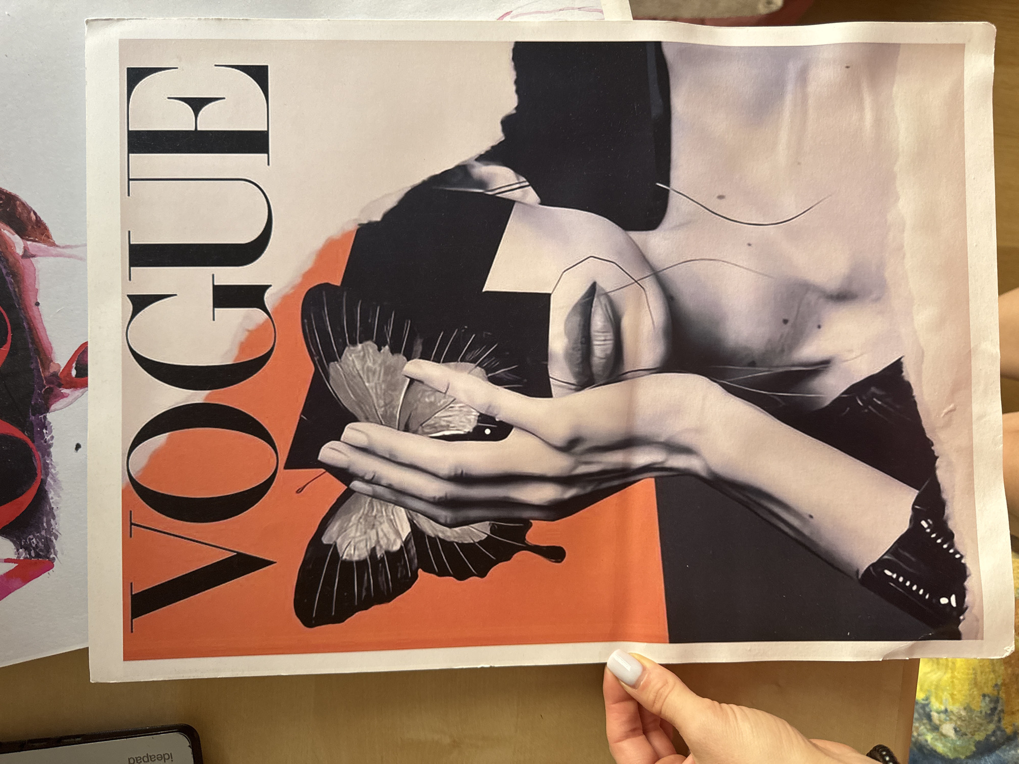 The image shows a Vogue magazine cover featuring a surreal collage of a woman's face, a butterfly, and abstract shapes. A hand with painted fingernails is visible on the left side of the frame, holding the edge of the magazine. The magazine is lying on a wooden surface, with another piece of artwork, possibly a painting or print, partially visible behind it at the top. The overall impression is of a creative workspace or a display of artistic materials. The location is not specified in the image or its surrounding context.