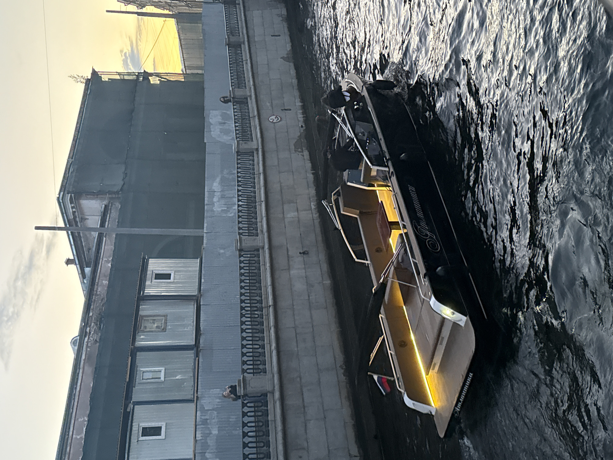 In Saint Petersburg, Russia, a dark-hulled boat with a wooden deck and illuminated interior is navigating a narrow canal. The boat, named "Атлантика" (Atlantika) in Cyrillic script, is moving from left to right. The water is choppy, reflecting the surrounding environment.
Along the canal's edge, a stone embankment is visible, topped with a metal railing. Behind the railing, a person is standing and observing the canal. Further up, a row of modular, corrugated metal structures with small windows are built against a larger, dark-clad building, which appears to be under construction or renovation, draped in dark netting. The sky above is a mix of muted yellow and grey clouds, suggesting either dawn or dusk. The overall scene depicts a typical urban waterway in Saint Petersburg during the transitional light of day.