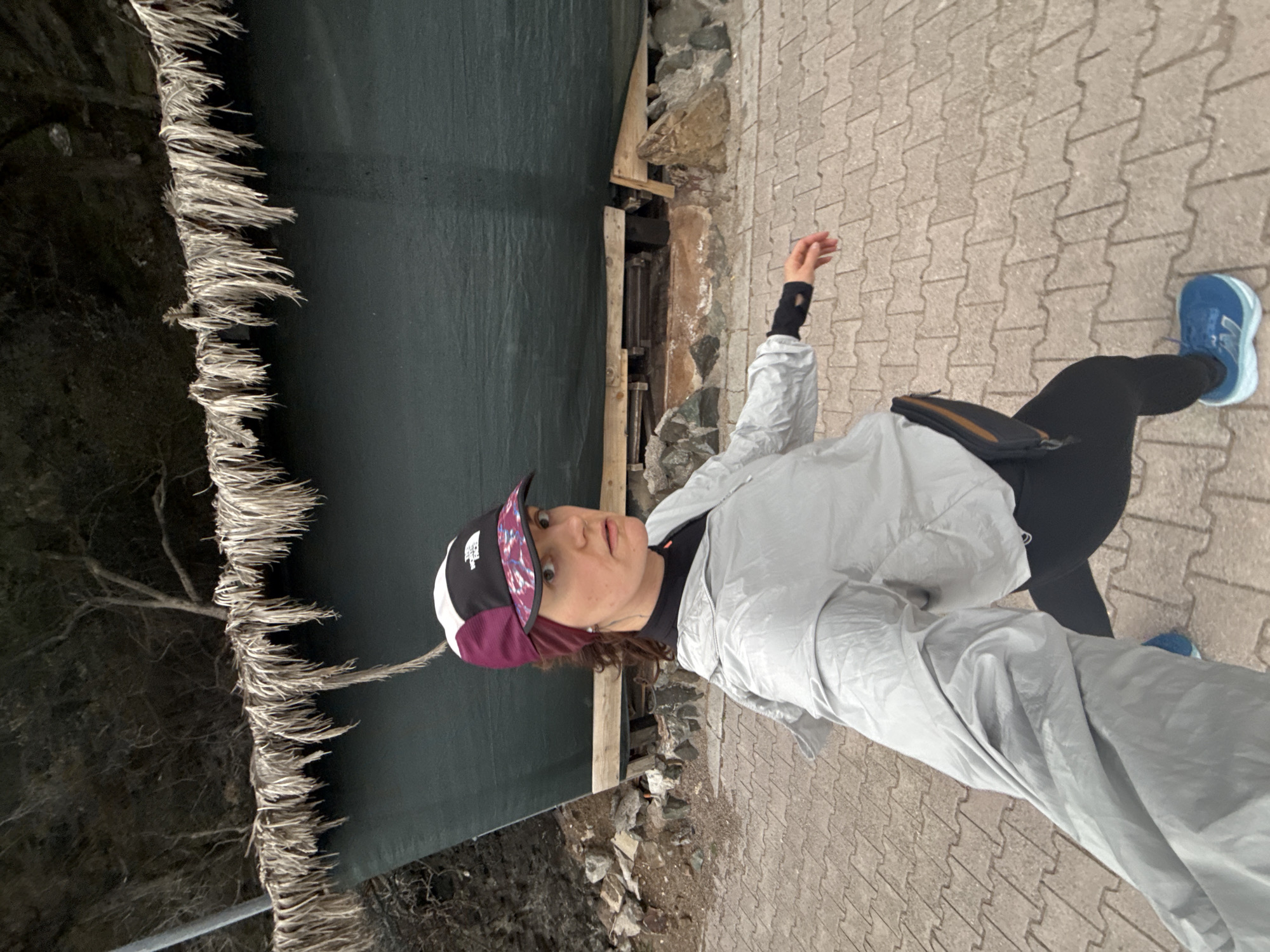A woman is standing in Boreti, Montenegro. She is wearing a white windbreaker, black leggings, and blue running shoes, and she has a black, white, and burgundy baseball cap on her head. She is standing on a paved walkway. Behind her is a body of water with dark green water. The water's edge is lined with dry vegetation. A retaining wall supports the path where the woman is standing.