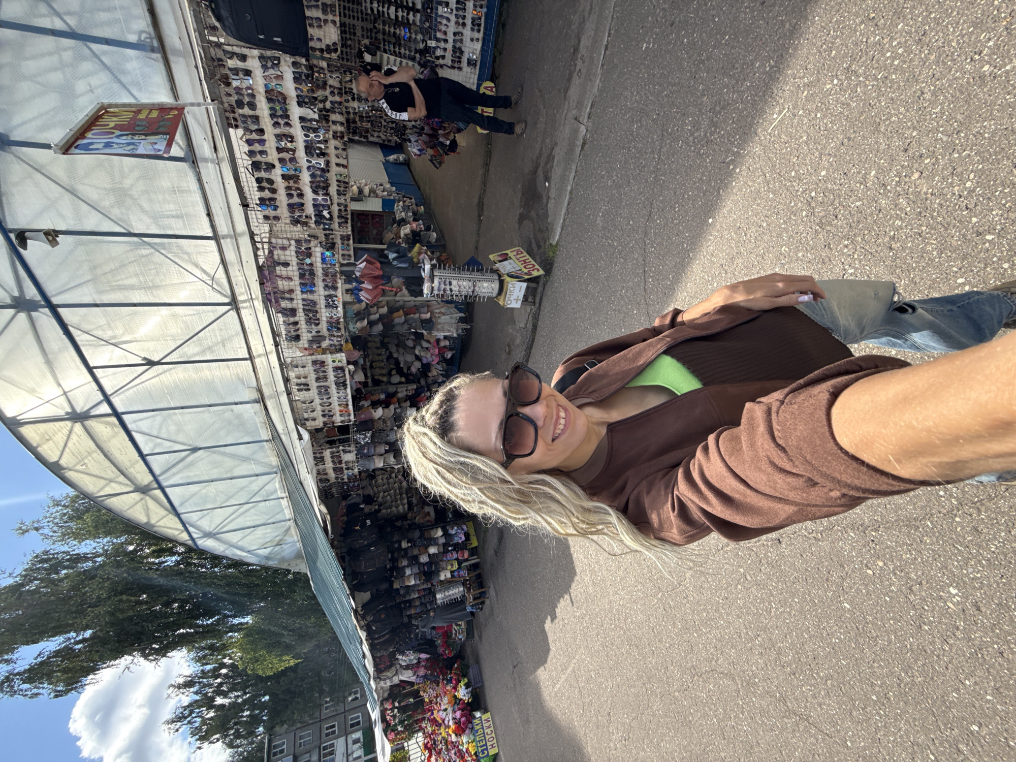 In Zhukovo, Russia, a young Caucasian woman with long blonde hair, wearing oversized sunglasses, a brown jacket, a green top, and ripped jeans, is taking a selfie in front of an outdoor market. The market stalls are covered by a translucent, arched roof and sell various items, including sunglasses, hats, and artificial flowers. A man in a black t-shirt and dark pants stands near a stall in the background. The ground is paved with asphalt. The sky is blue with scattered clouds, and trees and a multi-story building are visible behind the market.
