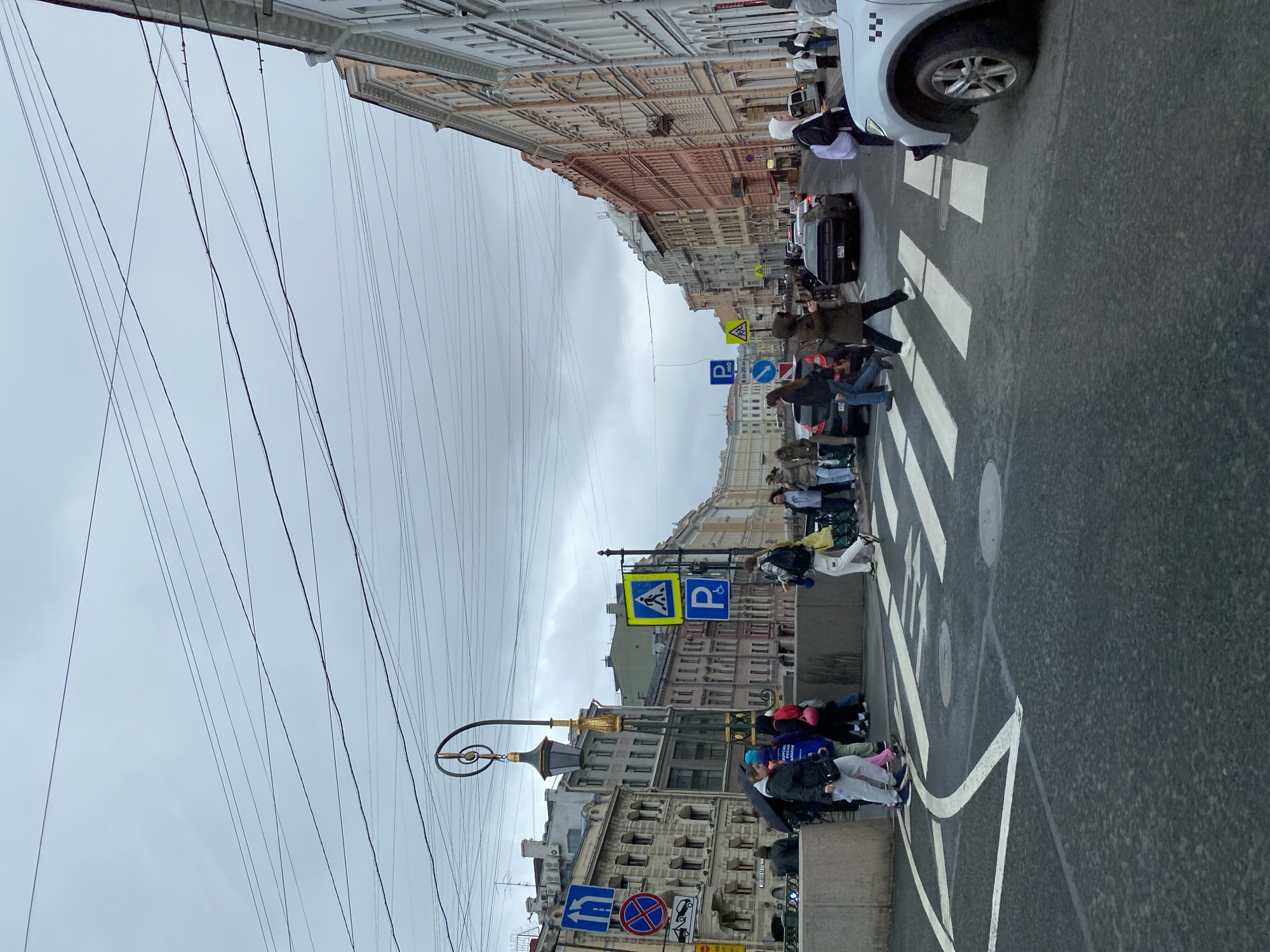 A street scene in an unidentified city shows pedestrians crossing a zebra-striped crosswalk. Traffic lights and road signs are visible, indicating pedestrian crossing and parking regulations. A white taxi is partially visible at the top right of the frame. Buildings line the street, their architecture suggests an urban environment. Power lines are strung across the sky above the street. The sky is overcast. People are walking and interacting at the crosswalk.