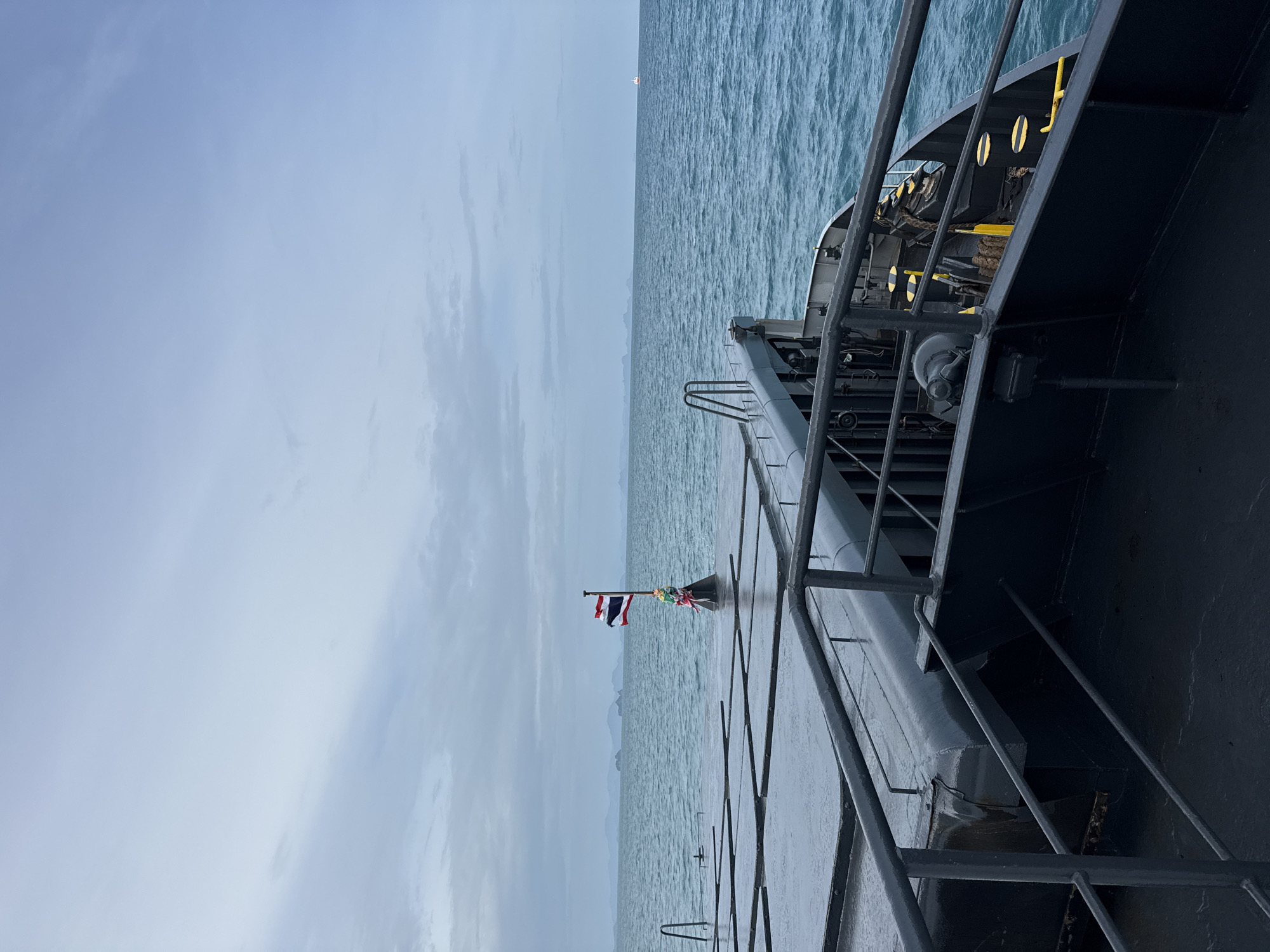 An outdoor, wide-angle, medium-shot photograph captures a view from the deck of a ferry as it travels across the ocean. The sky is overcast and a pale blue. The sea is a deep blue with moderate waves. In the distance, the silhouettes of islands are visible. A Thai flag flies from a flagpole on the ferry deck. The deck itself is dark and appears wet. The railing of the ferry runs along the right side of the frame. The image has a slightly muted color palette due to the cloudy conditions. The location is the Gulf of Thailand, near Ko Samui District, Thailand.