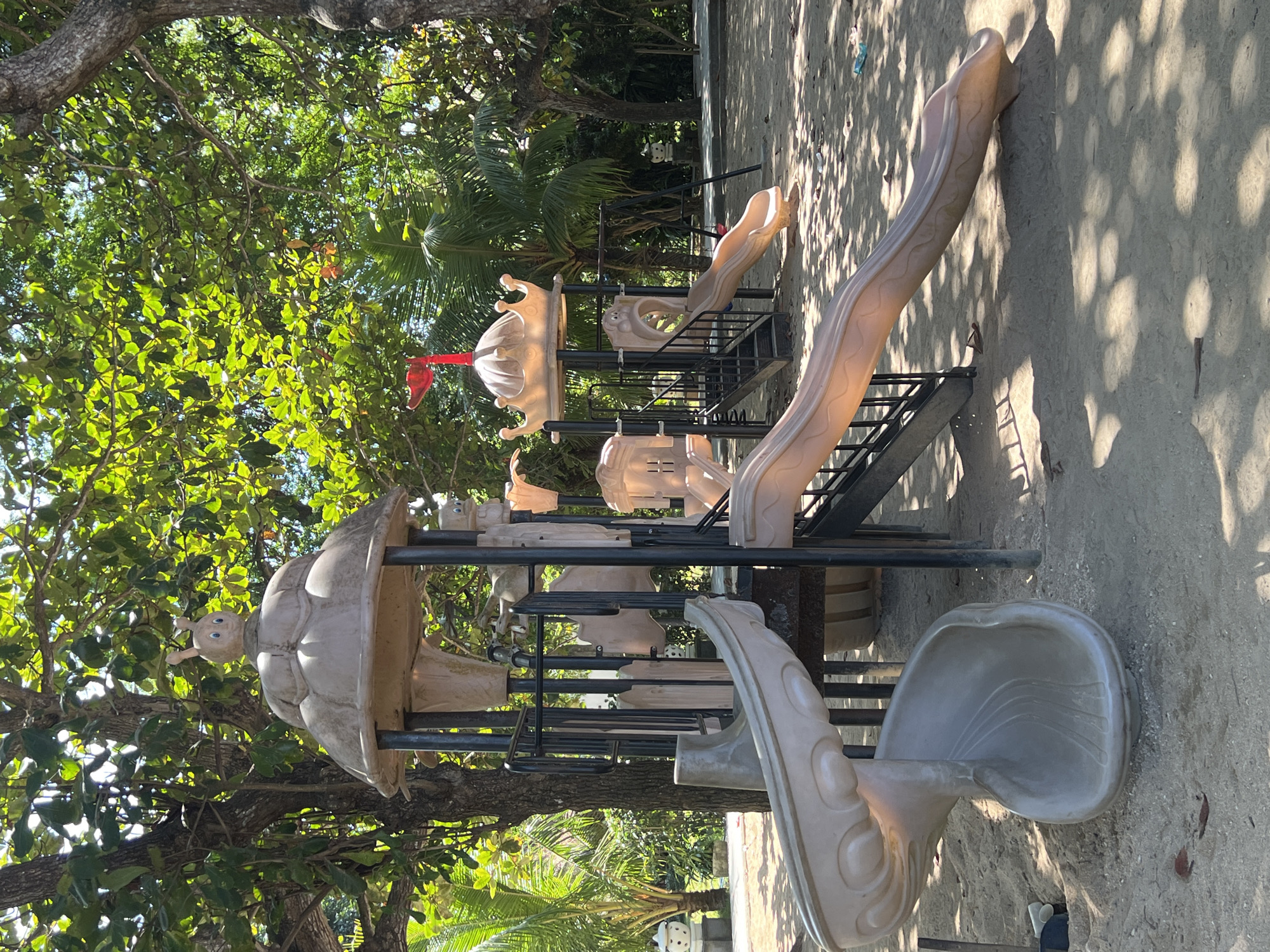 The image depicts an outdoor children's playground situated in Denpasar, Indonesia. The playground features a multi-level structure with several slides, including a curved gray slide on the left and a longer, lighter brown slide on the right. The structure is constructed from plastic and metal, with a whimsical design that includes a crown-like roof on one section, topped with a red flag. A cartoonish insect figure with big blue eyes is perched on top of the domed section. Stairs with black metal railings provide access to the elevated platforms. The playground is set on a sandy ground, with dappled sunlight filtering through the dense green foliage of mature trees that surround the area. Palm trees are also visible in the background, contributing to the tropical environment. Scattered on the sand are a few small pieces of debris. No people are present in the image.
