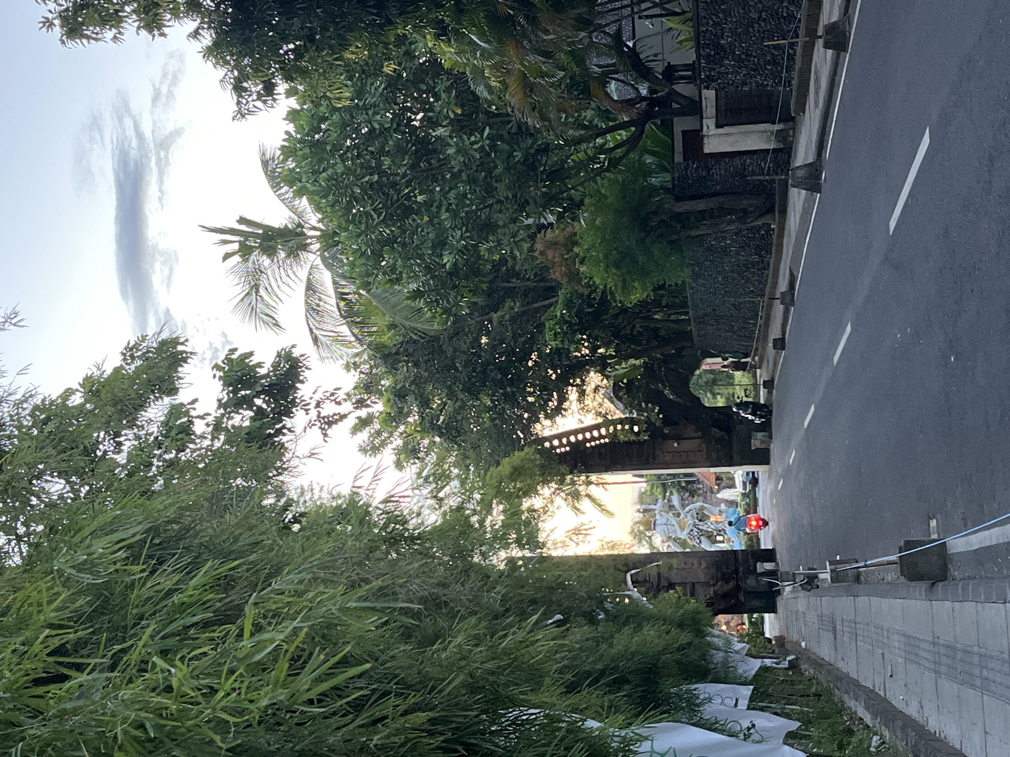 The image depicts a street scene in an unknown city in Indonesia. A paved road stretches into the distance, flanked by sidewalks on either side. Lush greenery, including bamboo and tropical trees, overhangs the street, creating a shaded canopy. Balinese stone walls and structures are visible along the roadside. A motorcycle with a red taillight is present on the road. In the distance, decorative statues can be seen near a traditional Balinese gate. The sky is partially cloudy, suggesting either early morning or late afternoon light.