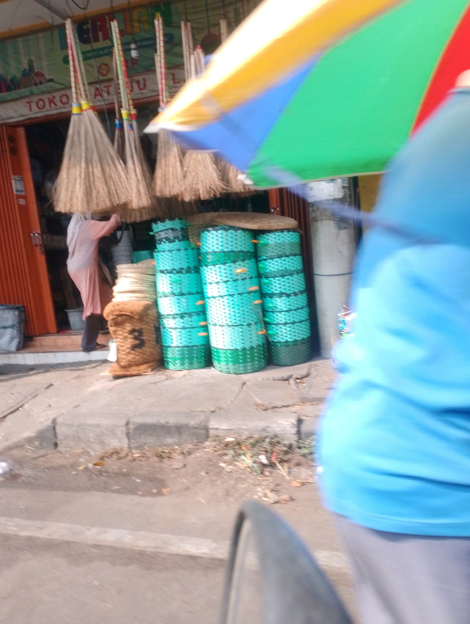 The image depicts a street scene in Probolinggo, Indonesia, likely outside a shop selling brooms. In the foreground, a person wearing a blue shirt and grey pants is partially visible, suggesting they are either walking or riding a vehicle. Behind them, a large, colorful umbrella with panels of yellow, blue, green, and red is open, partially obscuring the view of the shop.
The shop itself features an orange roll-up door. Hanging from the top of the shop entrance are numerous brooms, their straw ends facing downwards. A person, possibly a vendor, wearing a light pink hijab and a long pink garment, is seen inside the shop, arranging or attending to the brooms.
In front of the shop, stacked on the sidewalk, are several large, cylindrical baskets made of woven material, primarily green and turquoise. To the left of these baskets, a large, brown sack is leaning against the wall. The sidewalk is made of concrete, and there is debris scattered on the ground. The overall environment suggests a commercial area on a street in Probolinggo.