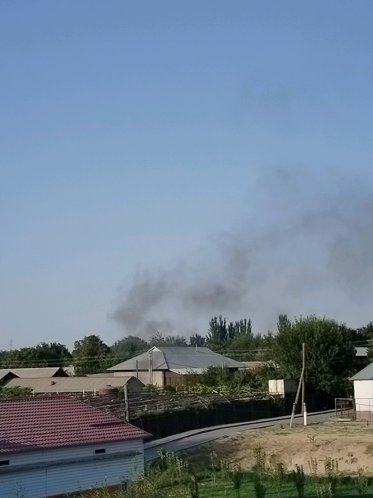 A plume of dark smoke is rising into the clear blue sky above a residential area in Kölkent, Kazakhstan. The smoke appears to be originating from behind a row of single-story buildings with corrugated metal roofs. Trees and vegetation are visible throughout the scene, interspersed with houses and fences. A road curves through the foreground, bordered by dry grass and a few small plants.