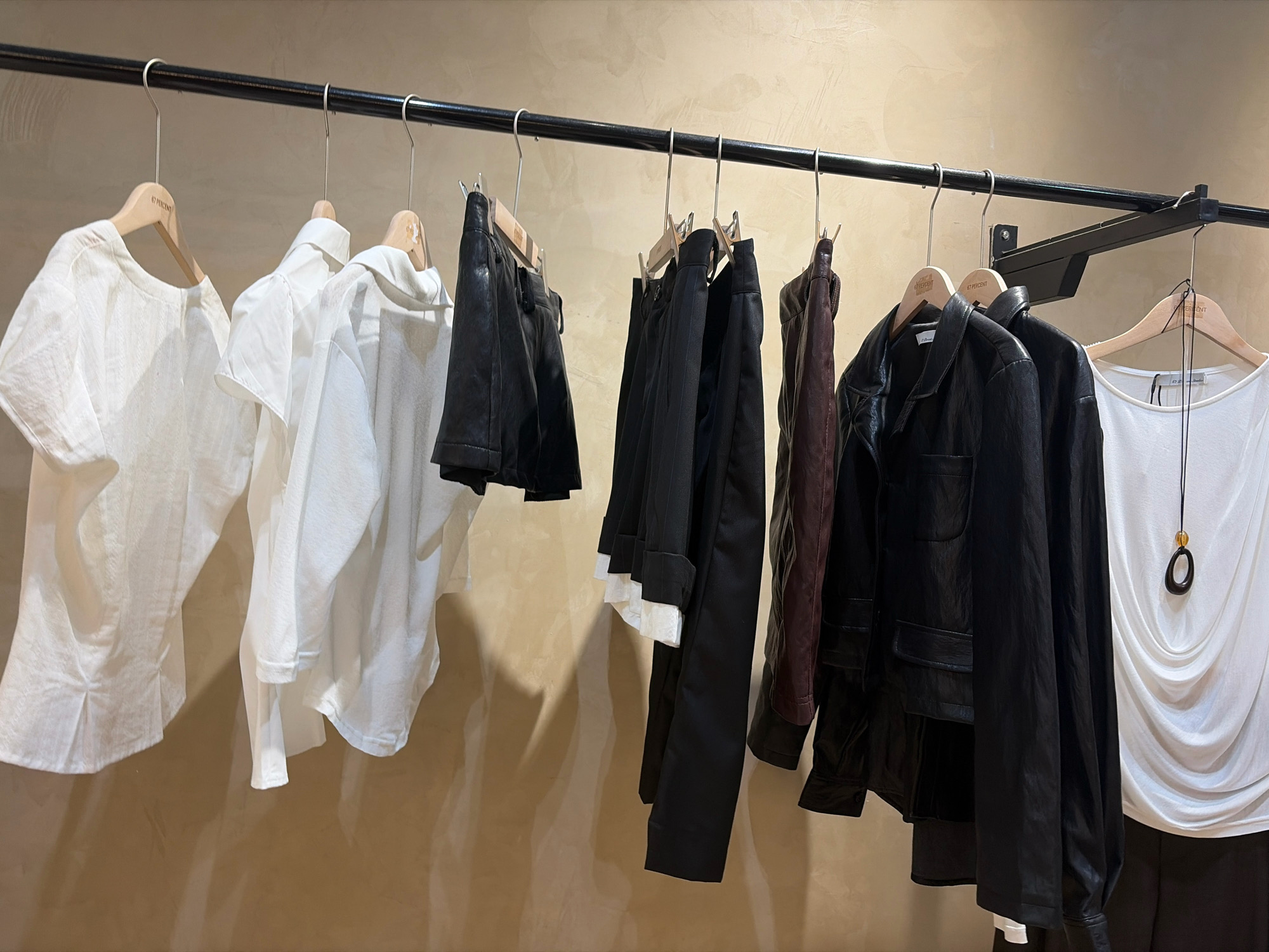 The image shows a clothing rack with various garments hanging on wooden and metal hangers against a textured beige wall. From left to right, the items include a white short-sleeved top with a textured fabric, a white collared shirt, another white shirt, two black leather-like shorts, black trousers, a dark reddish-brown jacket, a black jacket, and a white draped top with a necklace. The clothing appears to be displayed in a retail or studio setting. There are no people visible in the image. The lighting suggests an indoor environment.