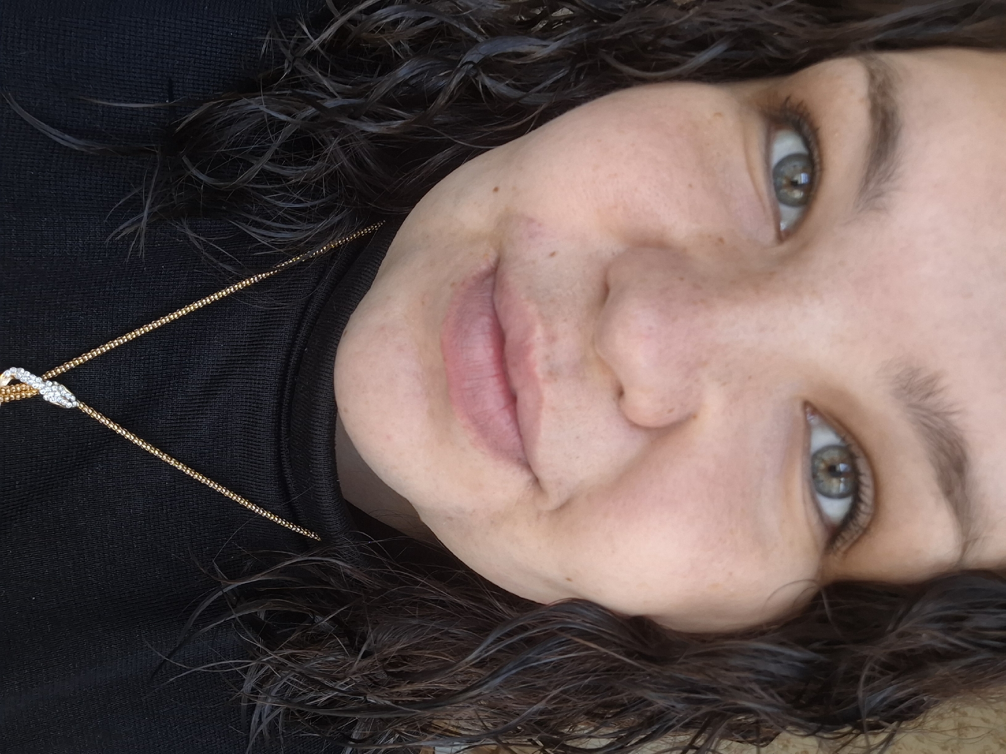 A close-up portrait features a person, likely female, with dark, curly hair that appears somewhat wet or styled. The head is tilted, presenting the image at an angle. The subject has a light complexion adorned with visible freckles and prominent green or hazel eyes. A soft, gentle smile plays on their lips. They are wearing a black high-neck or turtleneck top. Around their neck, a thin gold chain necklace with a small, intricate pendant, possibly featuring two intertwining elements, is visible against the dark fabric. The background is softly blurred, keeping the focus entirely on the subject's face. The lighting appears natural and diffused. No specific location or time of day cues are discernible from the image itself, other than the provided context of Марковский, Russia. There is no visible text.