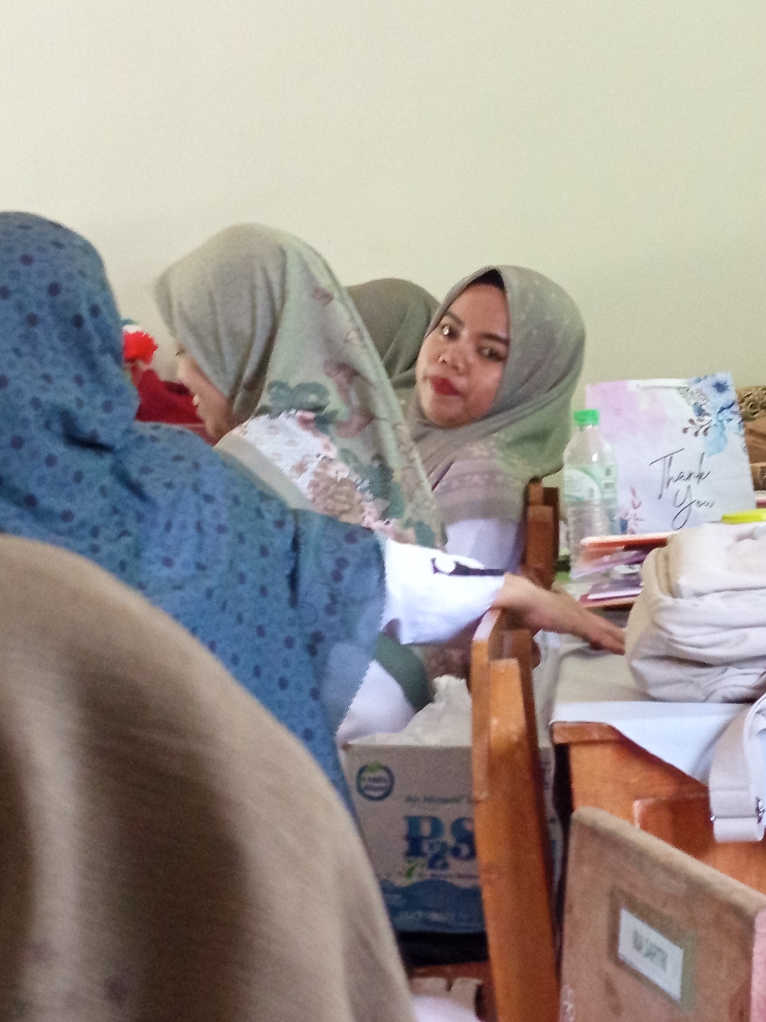 The image depicts a dimly lit indoor scene with three individuals, likely female, wearing hijabs. The central figure, facing towards the camera, has dark hair and is wearing a light green hijab and a red lipstick. To her left, another individual is partially visible, also wearing a hijab. A third individual is largely out of frame on the left, with a blue hijab featuring a dark polka-dot pattern.
On a table in the foreground, several objects are present: a bottle of water, a "Thank You" card with floral designs, a stack of books or papers, and a beige backpack or bag. A cardboard box with the text "PS" is also visible. The environment appears to be a classroom or a communal space, suggested by the tables and chairs. The image's quality is somewhat grainy, indicating it might have been taken with a lower-resolution camera or under suboptimal lighting conditions. The specific location in Lumutan, Indonesia, is not visually identifiable from the image content.