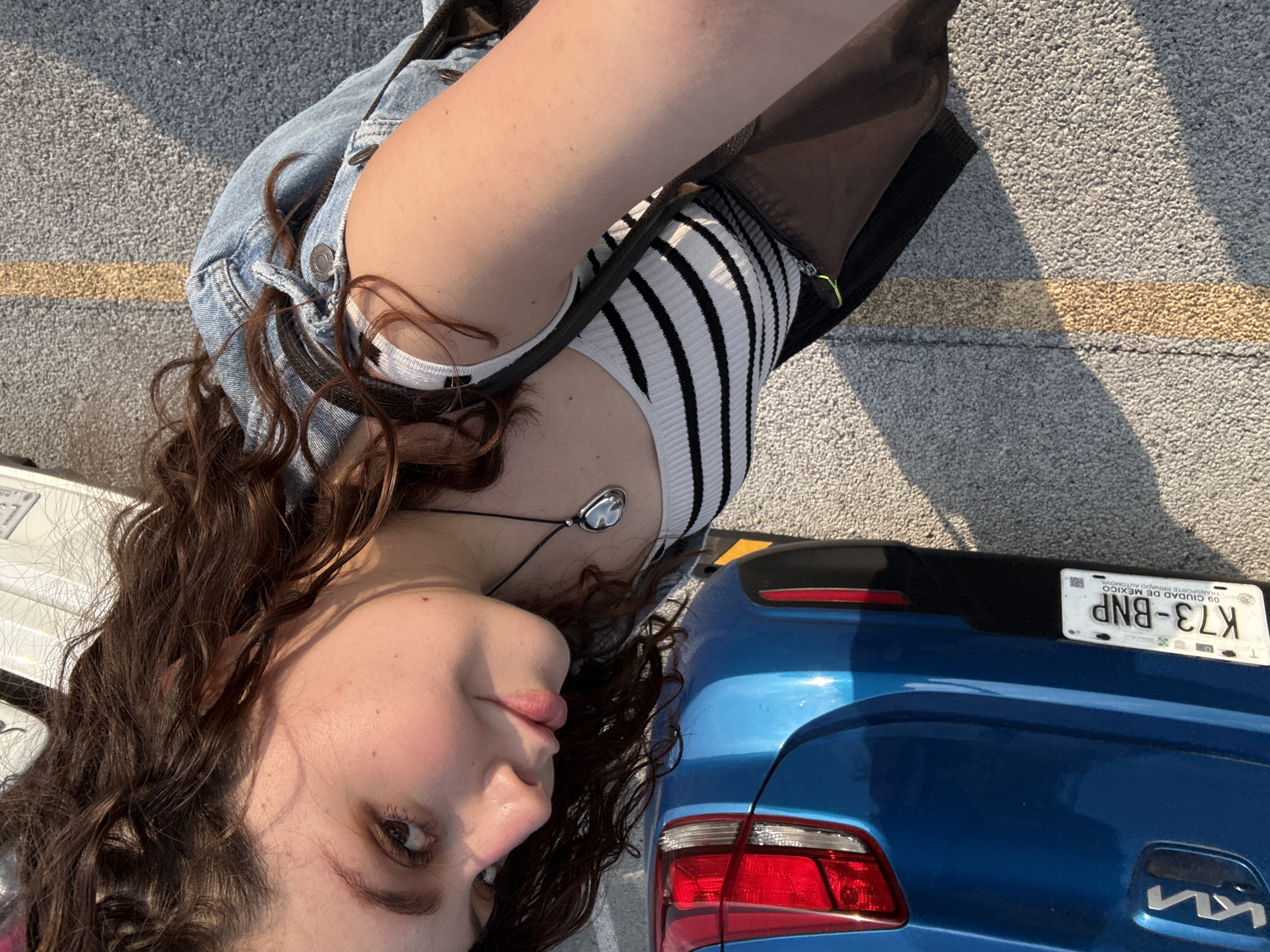 An overhead outdoor shot, rotated 90 degrees clockwise, captures a young person and the rear of a blue car on asphalt pavement. Bright sunlight illuminates the scene, casting distinct shadows.
The subject is a person with long, curly brown hair, wearing a white and black striped tank top layered under a light blue denim jacket. A necklace with a reflective, silver-colored oval pendant is visible around their neck. Their face, positioned towards the bottom left of the frame, is angled slightly away from the camera, revealing a neutral to subtly pensive expression. The person's left arm is bent, and the right arm is mostly out of frame.
To the right, the rear portion of a blue Kia car is visible. It features a red taillight assembly on the left side of its rear bumper. A black trim piece runs above the bumper, where a license plate is affixed. The license plate clearly displays "K73-BNP" and the text "CIUDAD DE MEXICO", confirming the location as Mexico City, Mexico. Below the license plate, the "KIA" logo is visible on the car's tailgate.
The ground is composed of grey asphalt, with a faint yellow parking line running horizontally across the middle of the frame. The strong sunlight indicates it is daytime. The overall impression is a casual, sunlit outdoor moment.