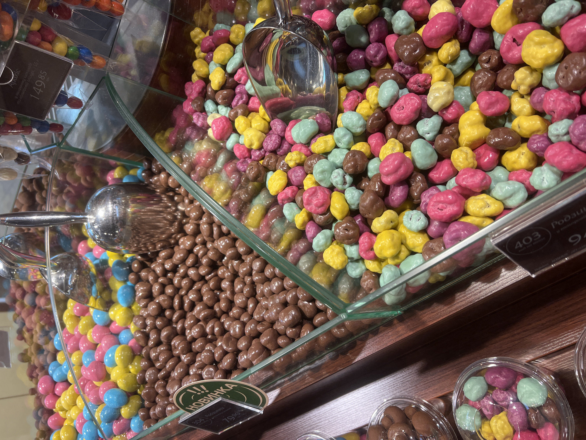 This image depicts a display of colorful candies in a store, likely a confectionary or grocery store, located in Odesa, Ukraine. The candies are presented in glass containers, with several scoops for customers to serve themselves. The dominant colors of the candies are pink, yellow, blue, and brown. In the foreground, there are clear plastic containers filled with a mix of pink, yellow, green, and brown candies. Behind these, larger glass displays showcase more of the same candies, some in bulk and others in pre-packaged bags with labels. One label is partially visible and appears to be in Cyrillic script. The overall environment suggests a retail setting focused on sweets.