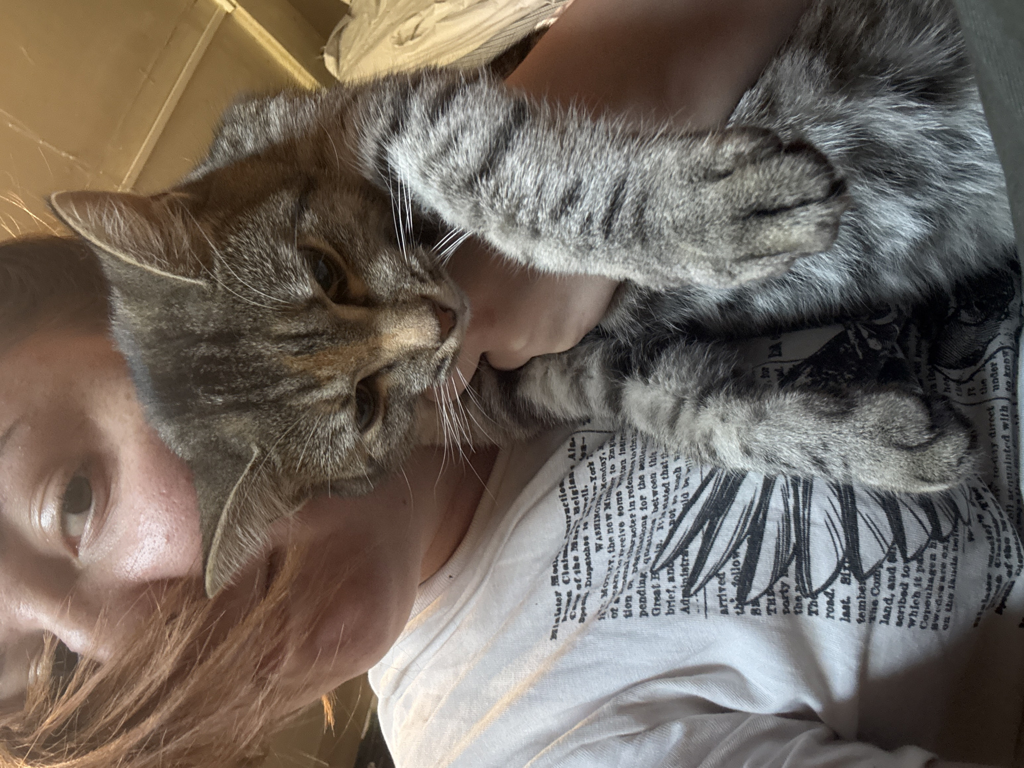 A person is holding a tabby cat. The person has red hair and is wearing a white t-shirt with black print. The cat is mostly gray with black stripes. The location is not discernible from the image.