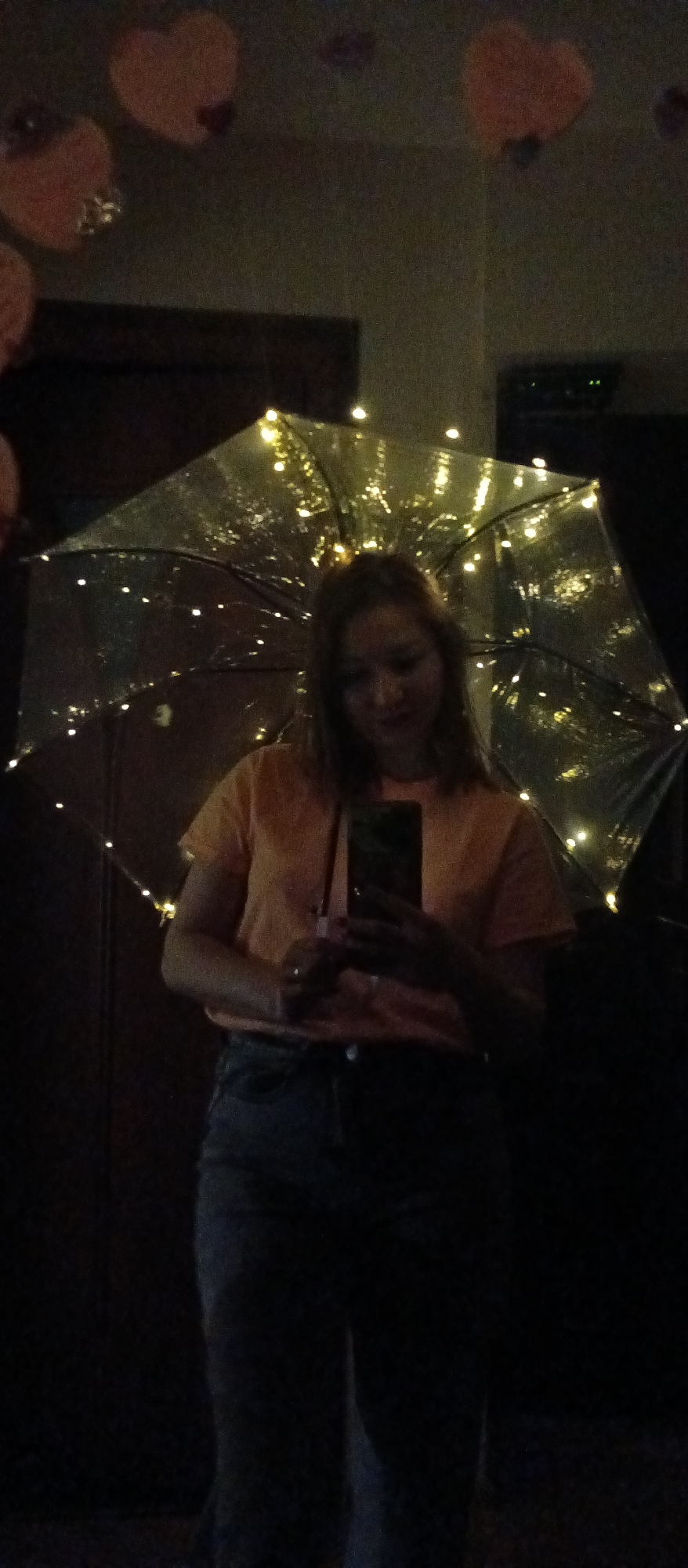 The image depicts a person, likely a young woman, standing indoors in a dimly lit setting. She is positioned centrally, holding a smartphone with both hands, suggesting she might be taking a selfie or viewing content on the screen. Her face is partially obscured by the phone and the low light, but she appears to be looking down at the device.
She is wearing a short-sleeved, light orange or peach-colored t-shirt and dark-colored jeans or pants. Behind and above her, she holds a large, transparent umbrella that is illuminated by numerous small, warm-toned string lights wrapped around its frame. These lights are the primary source of illumination in the scene, casting a warm glow and creating a festive or magical ambiance.
In the upper background, several blurred, heart-shaped objects, possibly balloons or cutouts, are visible, adding to a decorative theme. The overall background is very dark and indistinct, appearing to be a plain wall or door, which further emphasizes the illuminated umbrella and the subject. The photograph has a grainy texture, characteristic of low-light photography. There is no visible text in the image. The scene conveys a sense of a private moment or a celebration in a cozy, nocturnal indoor environment.