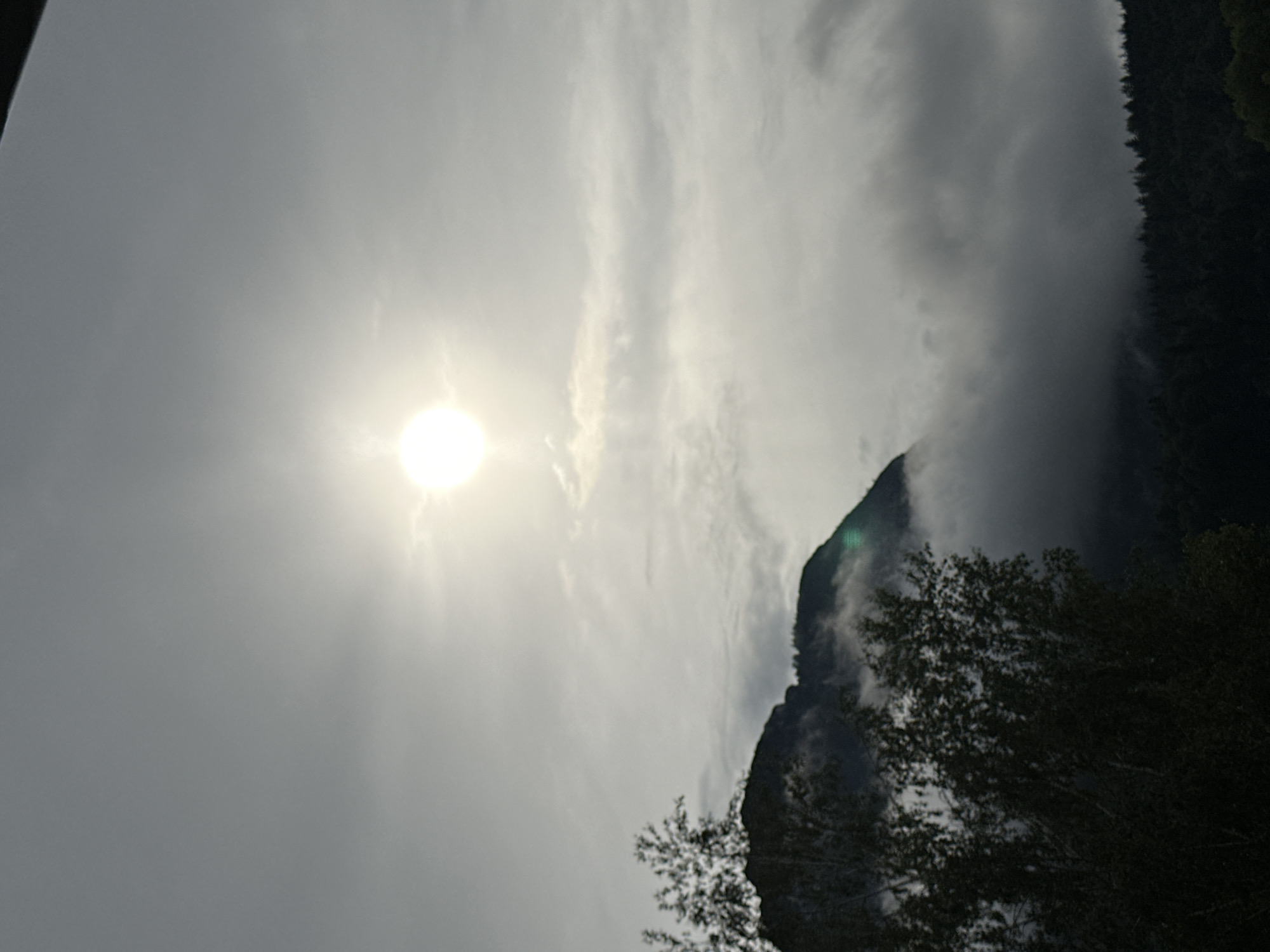 This media file depicts a scenic mountain landscape with the sun visible in the sky. The scene is dominated by a bright, hazy sun in the upper left quadrant, casting a strong glow through scattered clouds. A dark, tree-covered mountain range occupies the right side of the image, with fog or clouds partially obscuring its lower slopes. The foreground features silhouetted trees, suggesting a natural, possibly wild, environment.
The time of day appears to be daytime, given the sun's prominence, though the haze and cloud cover make it difficult to ascertain the exact hour. The weather seems partly cloudy with some atmospheric obscurity. The presence of mountains and dense forest strongly suggests a mountainous region. The overall mood conveyed is one of natural grandeur and perhaps a touch of mystery due to the obscured mountain. There is no visible text in the image. The provided location context of Garmisch-Partenkirchen, Germany, aligns with the visual elements of a European alpine setting.