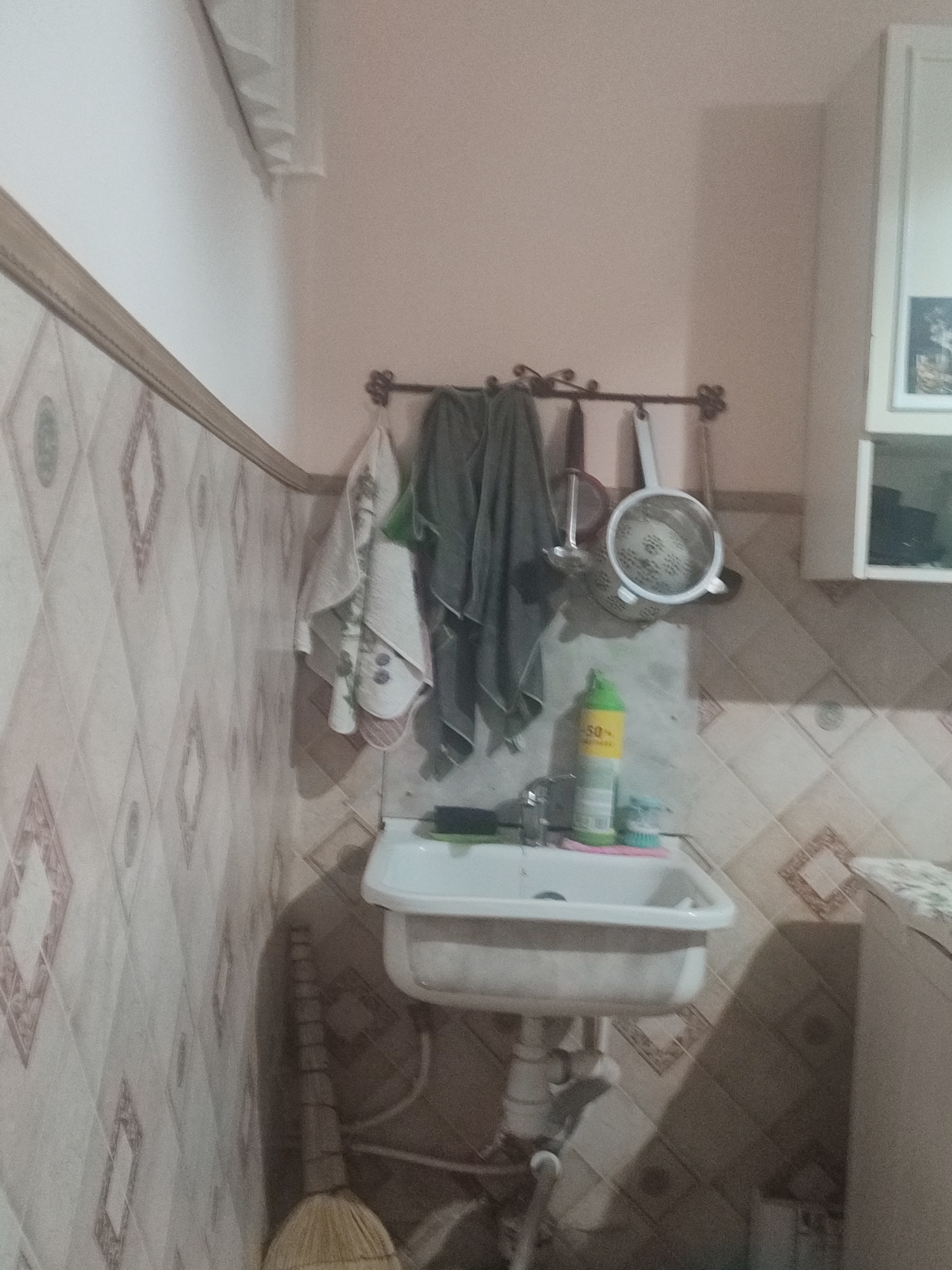 This is a medium-shot, indoor photograph of a small sink area, possibly a kitchen or utility room, with tiled walls. In the center of the image is a white sink with a chrome faucet. To the left of the sink, a broom with a straw head stands upright against the wall. Above the sink, a metal rack holds several kitchen items, including a colander, tongs, and what appear to be folded towels or cloths. A green and yellow bottle of cleaning spray stands on the sink's edge. The walls are covered in light-colored tiles with a diamond pattern. The floor is not clearly visible. The lighting is dim, casting shadows in the room. The image does not contain any information that would allow for its localization to Chernivtsi, Ukraine, or any other specific city.