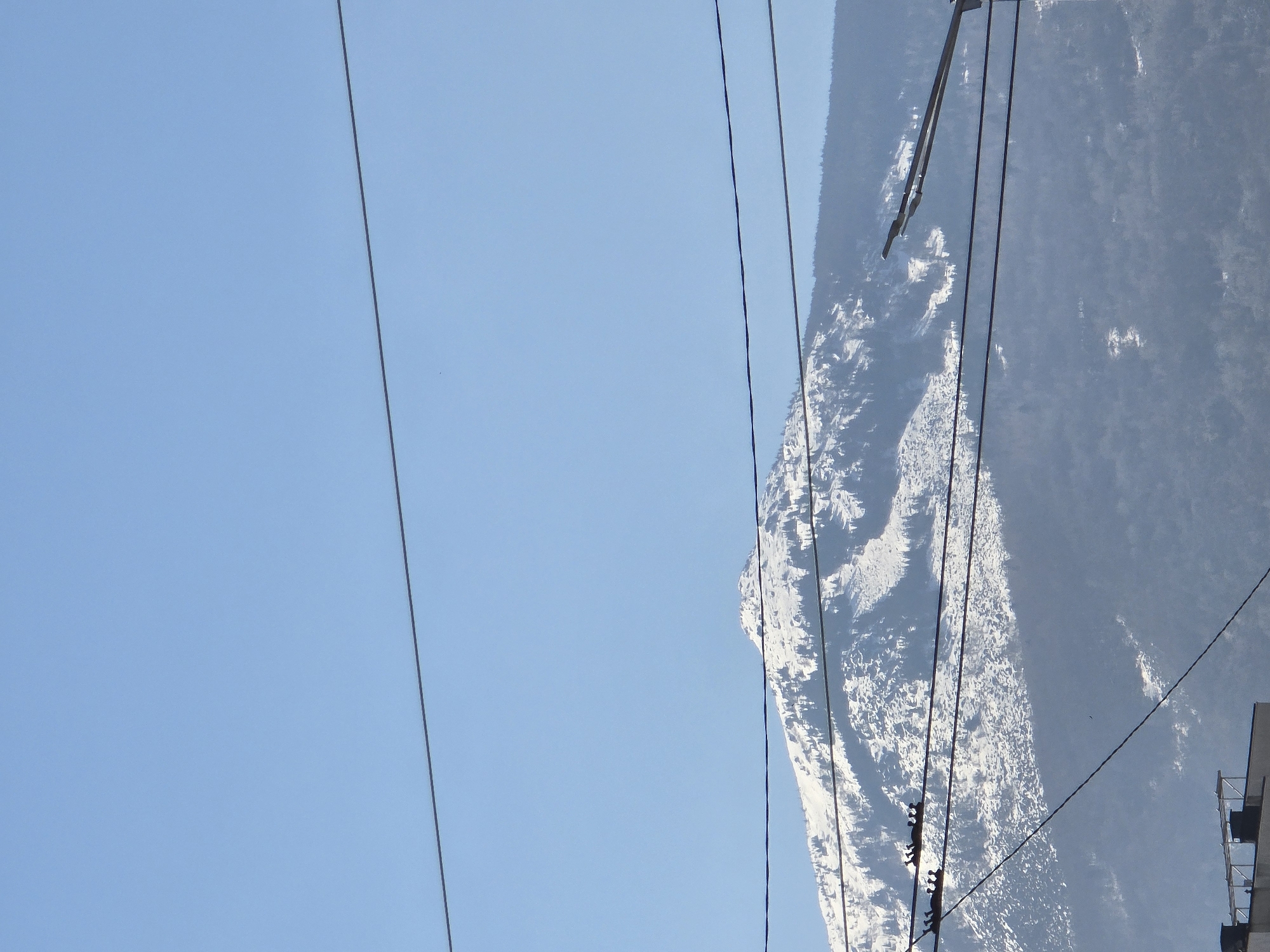 The image shows a mountain covered in snow, with power lines running across the view. The sky is clear and blue. The location could be a mountainous area near Sofia, Bulgaria, given the terrain and appearance of the landscape. The image includes a mountainous environment, snow, power lines, and a clear blue sky. There is a hint of a building at the bottom edge of the image.