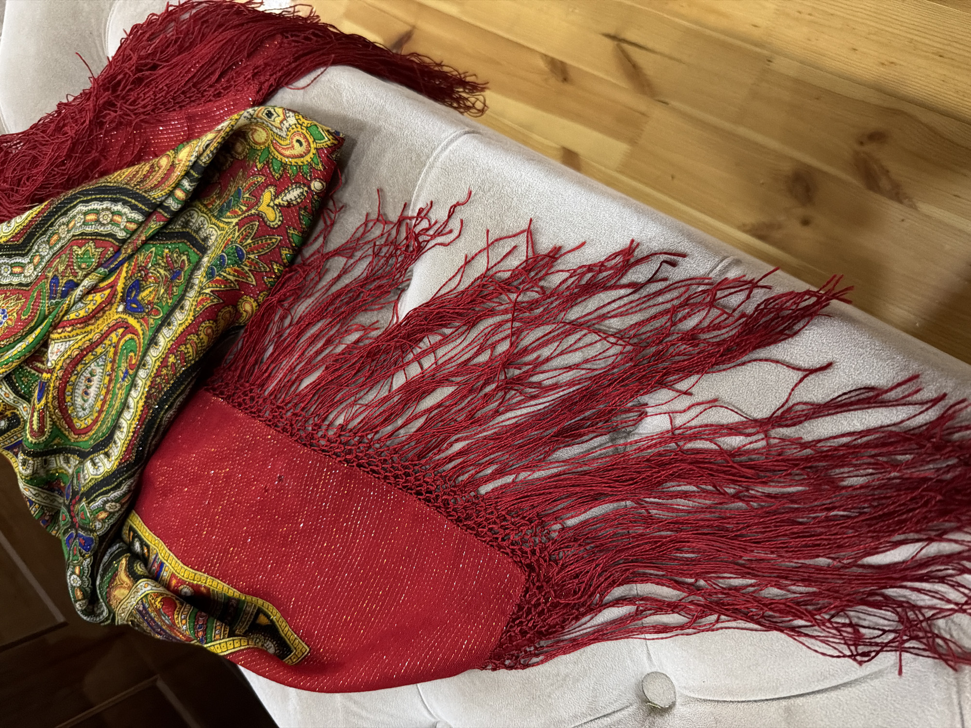 A close-up, high-angle shot shows a red scarf with a gold border and intricate paisley patterns in red, yellow, green, blue, and black, adorned with red fringe, draped over a light grey, tufted ottoman. The ottoman is positioned on a light-colored wooden floor. The background is blurred and shows a hint of wooden furniture. The scene appears to be indoors in a domestic setting. There are no people or any indications of Sabirabad, Azerbaijan.
