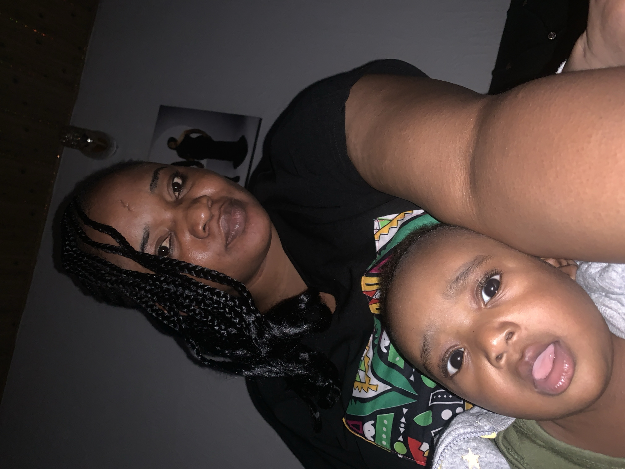 The media file shows a close-up, selfie-style shot of two people, an adult and a child, in what appears to be an indoor setting. The adult, positioned in the foreground and to the left, has braided hair and is looking towards the camera with an open mouth. The child, in the foreground and to the right, is also looking towards the camera with their tongue sticking out slightly. Both subjects have dark skin tones. The adult is wearing a black t-shirt, and the child is wearing a grey hooded jacket over a green shirt with a colorful patterned fabric visible underneath.
The background is dimly lit. To the left, a light source is visible, possibly a lamp, and a wooden surface with a textured pattern. Behind the adult's head, a framed image is partially visible, depicting a silhouetted figure. The overall lighting suggests it could be nighttime or a poorly lit room. The subjects' expressions are playful and informal, indicating a casual moment. No visible text is present in the media file. The context provided places the scene in Barakin Akawo, Nigeria.