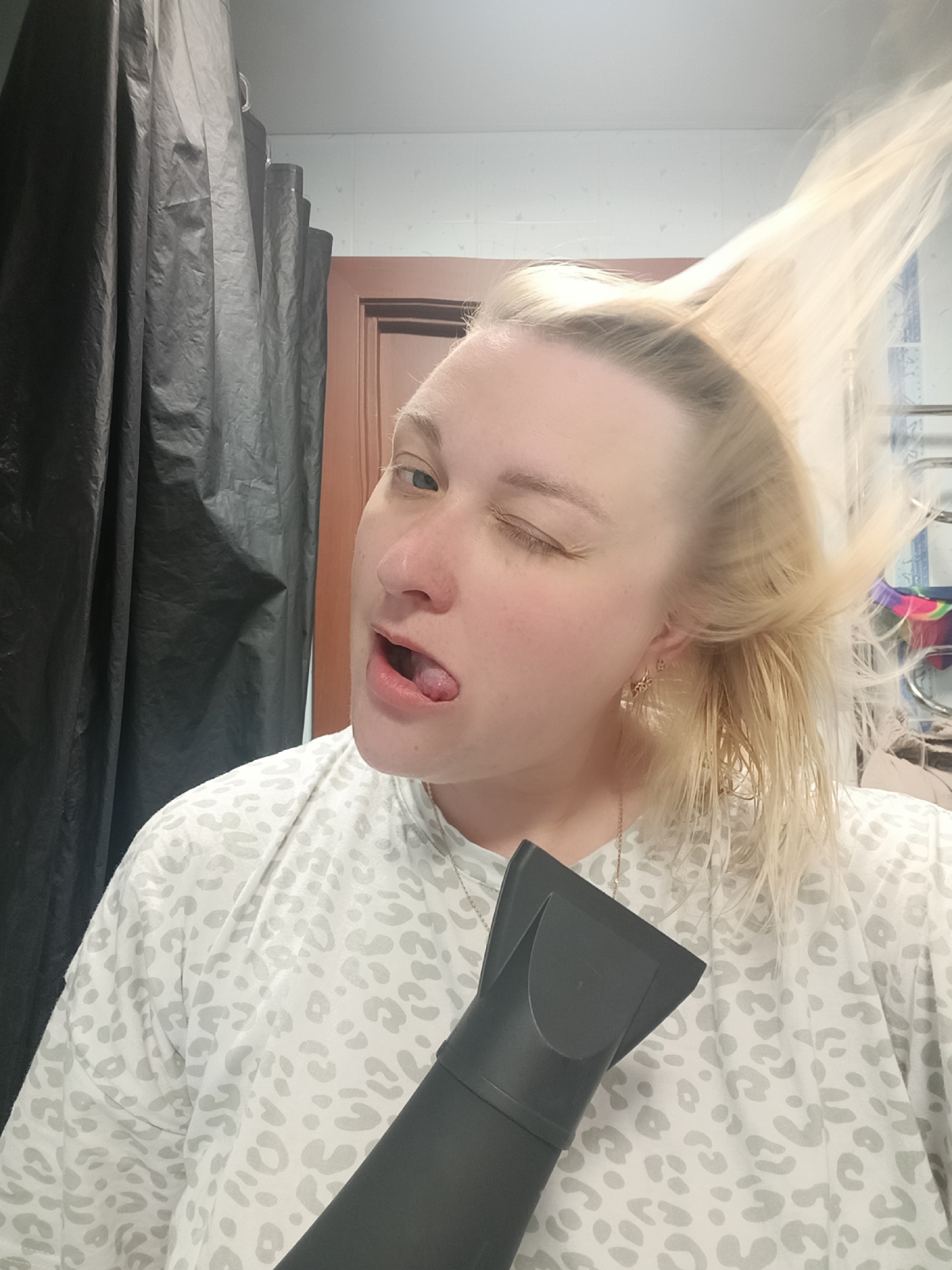 A woman with blonde hair, wearing a white and grey patterned shirt and gold earrings, is holding a black hairdryer with a nozzle attachment close to the camera. She is winking one eye and sticking out her tongue. Her hair is partially wet and styled upwards, as if being blown by the hairdryer. Behind her is a dark shower curtain, a wooden door, and a tiled wall. The location is likely a bathroom. The information about the city of Chaikovsky, Russia is likely extraneous and not visually discernible from the image.