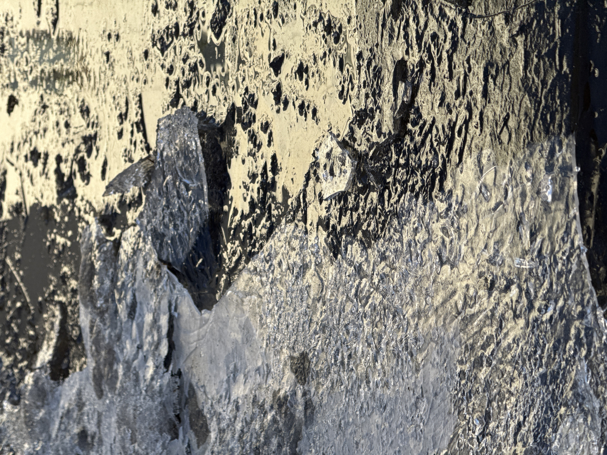 The image depicts a close-up view of ice formations on a surface. The surface appears rough and uneven with patterns of dark and light areas, potentially indicating dirt or debris embedded in the ice. There are distinct ice crystals and frozen textures. Given the mention of the city of Chaikovsky, Russia, it is plausible that this photograph was taken in this location, known for its cold climate and icy conditions.