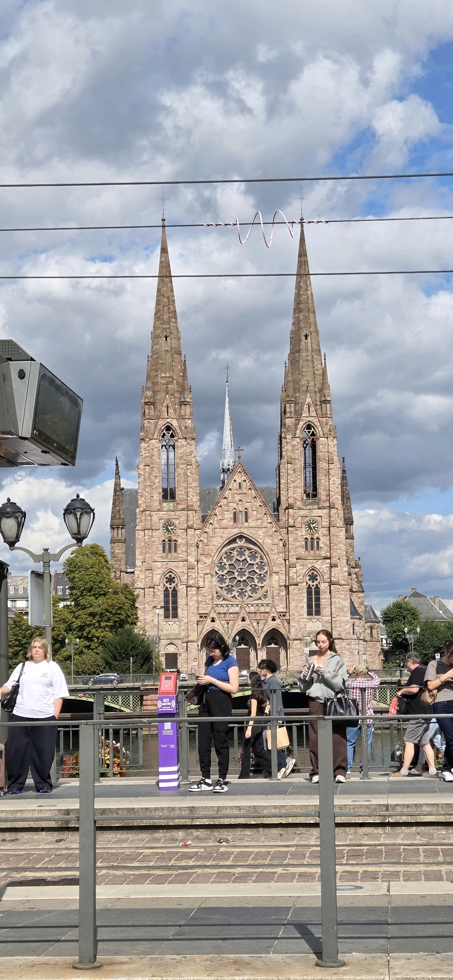The image shows a street scene in Strasbourg, France, featuring a prominent cathedral with twin spires. In the foreground, a group of people are standing on a platform, possibly a tram stop, with a metal railing. The people are diverse in age and gender, and some are engaged with their mobile phones. The cathedral, made of reddish stone, has large arched windows and intricate architectural details. The sky is partly cloudy, with the sun casting shadows. The overall atmosphere appears to be a typical urban day with people going about their business.