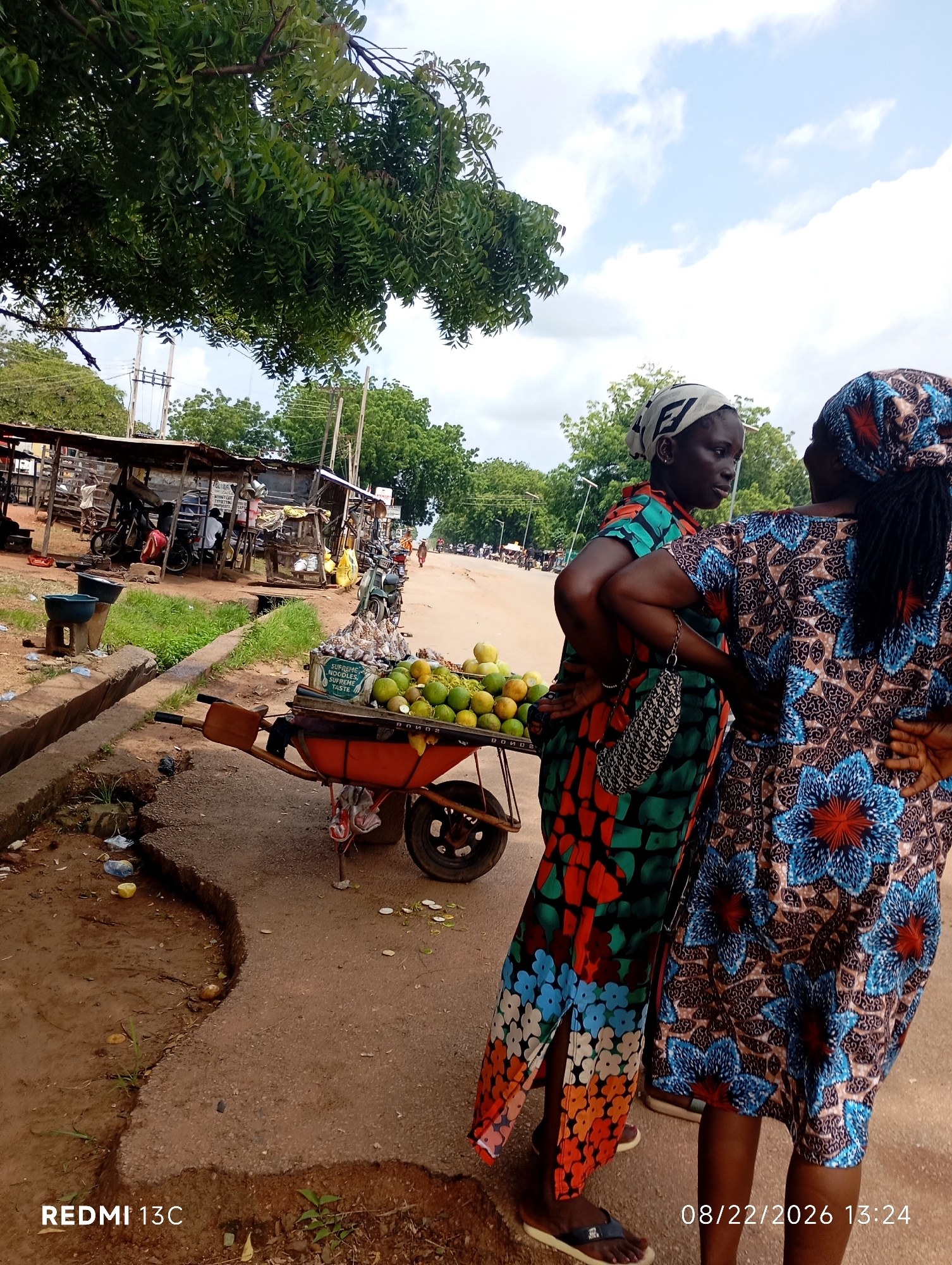 The image depicts a street scene in Shendam, Nigeria, on August 22, 2026, at 13:24. Two women are the primary subjects, standing near a fruit cart. The woman on the left wears a green and red patterned dress and a white and black patterned headscarf. She holds a patterned purse on her left arm. The woman on the right wears a blue, brown, and red patterned dress and a blue and red patterned headscarf. They appear to be conversing. In front of them is a red wheelbarrow filled with green fruits, possibly oranges, and some packaged goods with the text "SUPREME NOODLES" visible. The background shows market stalls with various items, motorcycles, and trees under a cloudy sky. The ground is a dirt and paved road with some debris.
