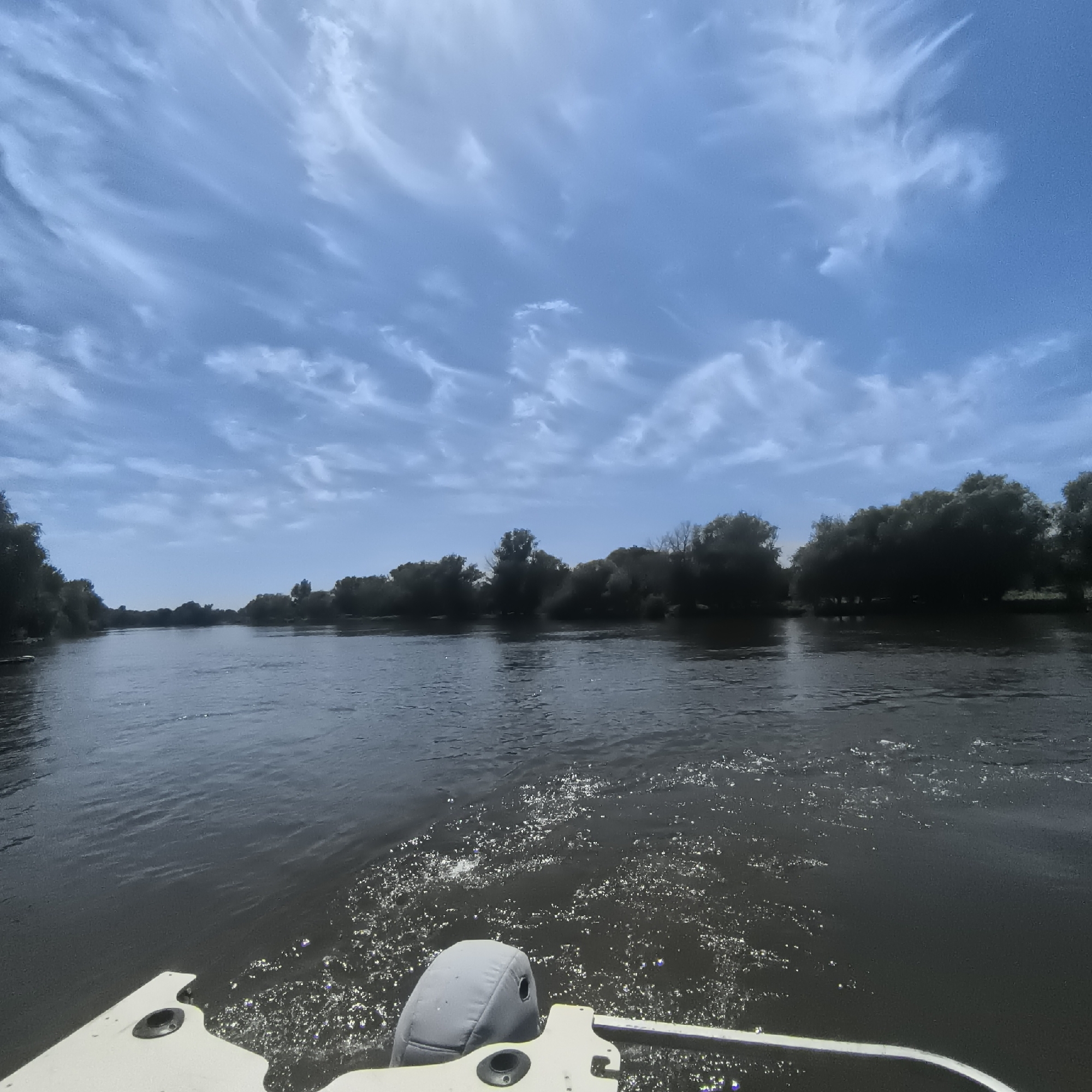 The image shows a view from a boat moving on a river. The boat's outboard motor cover is visible in the lower right corner. The river is dark and reflects the sky, which is mostly blue with wispy white clouds. The riverbanks are lined with dense trees. The scene is likely outdoors in a natural environment, possibly a rural area. The location is identified as Malyy Baklaniy, Russia.