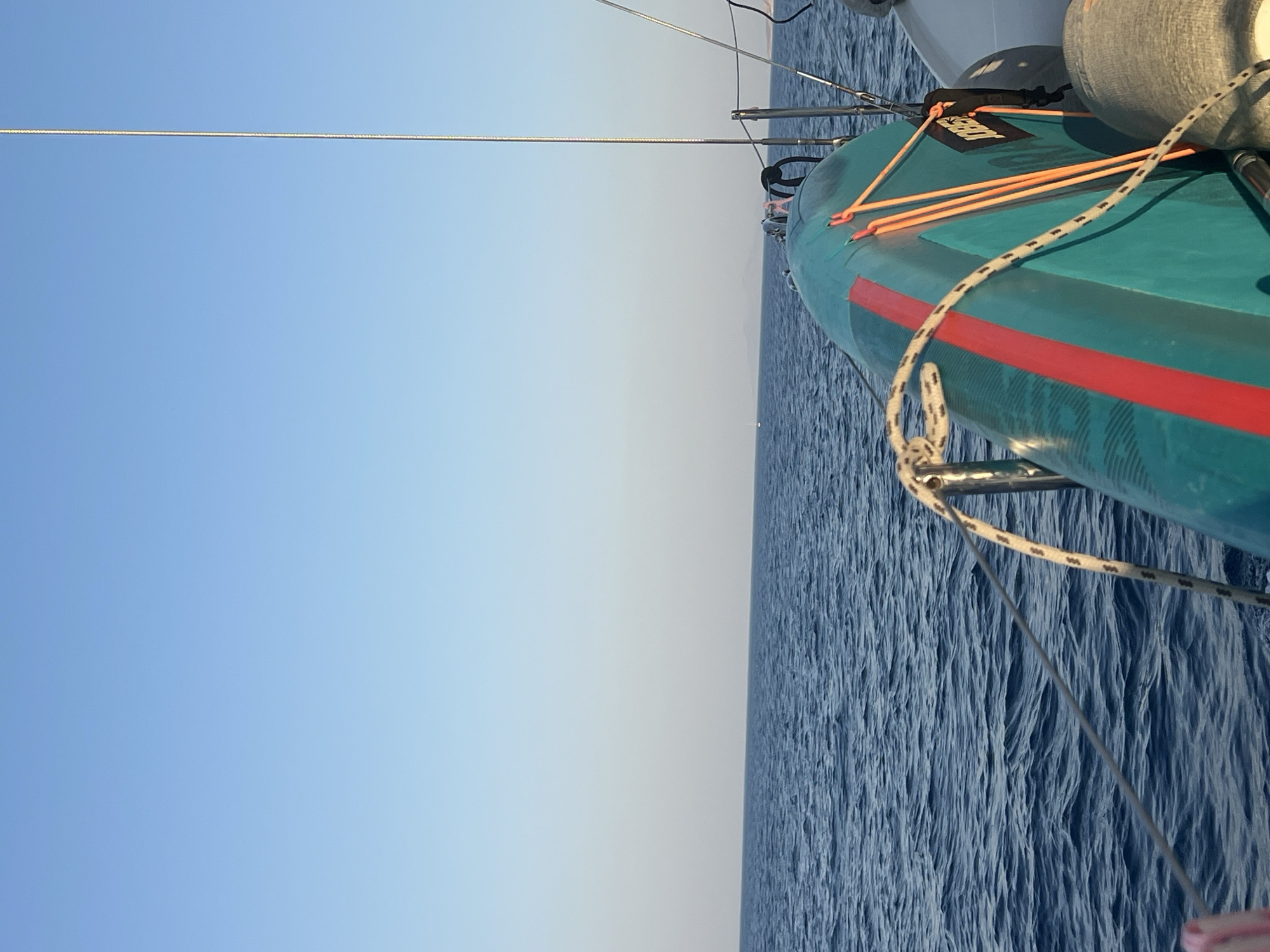 The image is a wide-angle shot taken from a boat on the water. The sky is clear and blue, with a slight gradient towards white on the horizon. The ocean is a deep blue with gentle waves. In the foreground, on the right side of the frame, part of the boat is visible, including a turquoise surfboard with a red stripe and orange ropes securing it. A coiled rope is also visible in the foreground. In the distance, faint landmasses are visible on the horizon. The overall scene suggests a calm day at sea.
