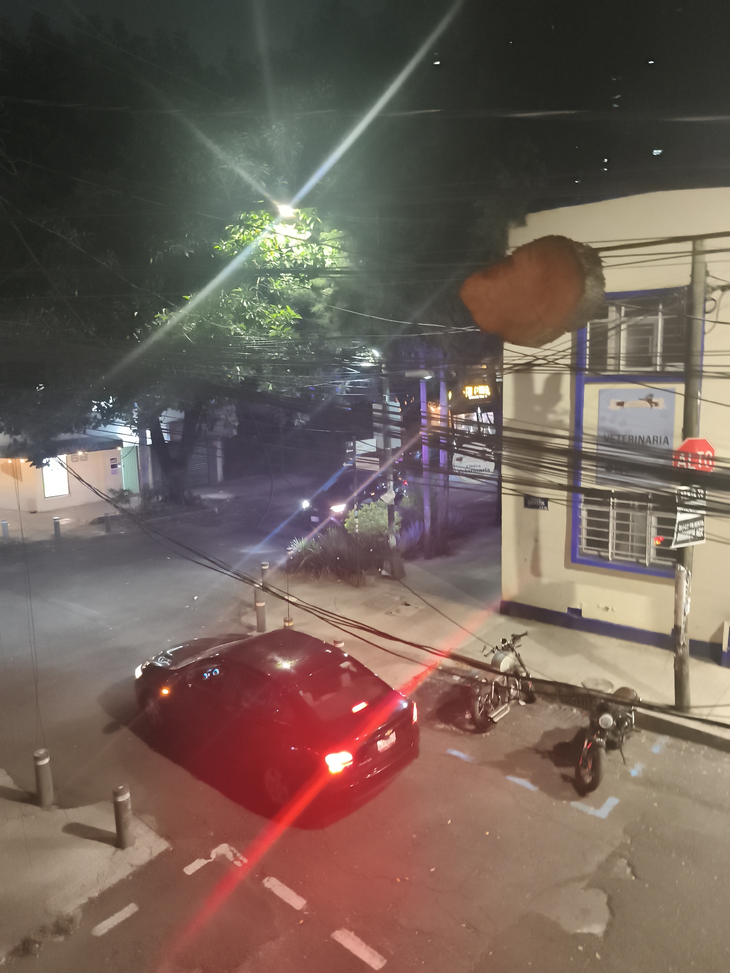 At night, the image depicts a street scene in Mexico City, Mexico. A red car with its taillights illuminated is parked on the left side of the frame. Next to the car are two motorcycles, also parked. Utility poles and wires crisscross above the scene. A building with the word "VETERINARIA" (Veterinary) and a blue-framed sign is on the right side of the frame. A street lamp provides illumination. Trees are visible in the background.