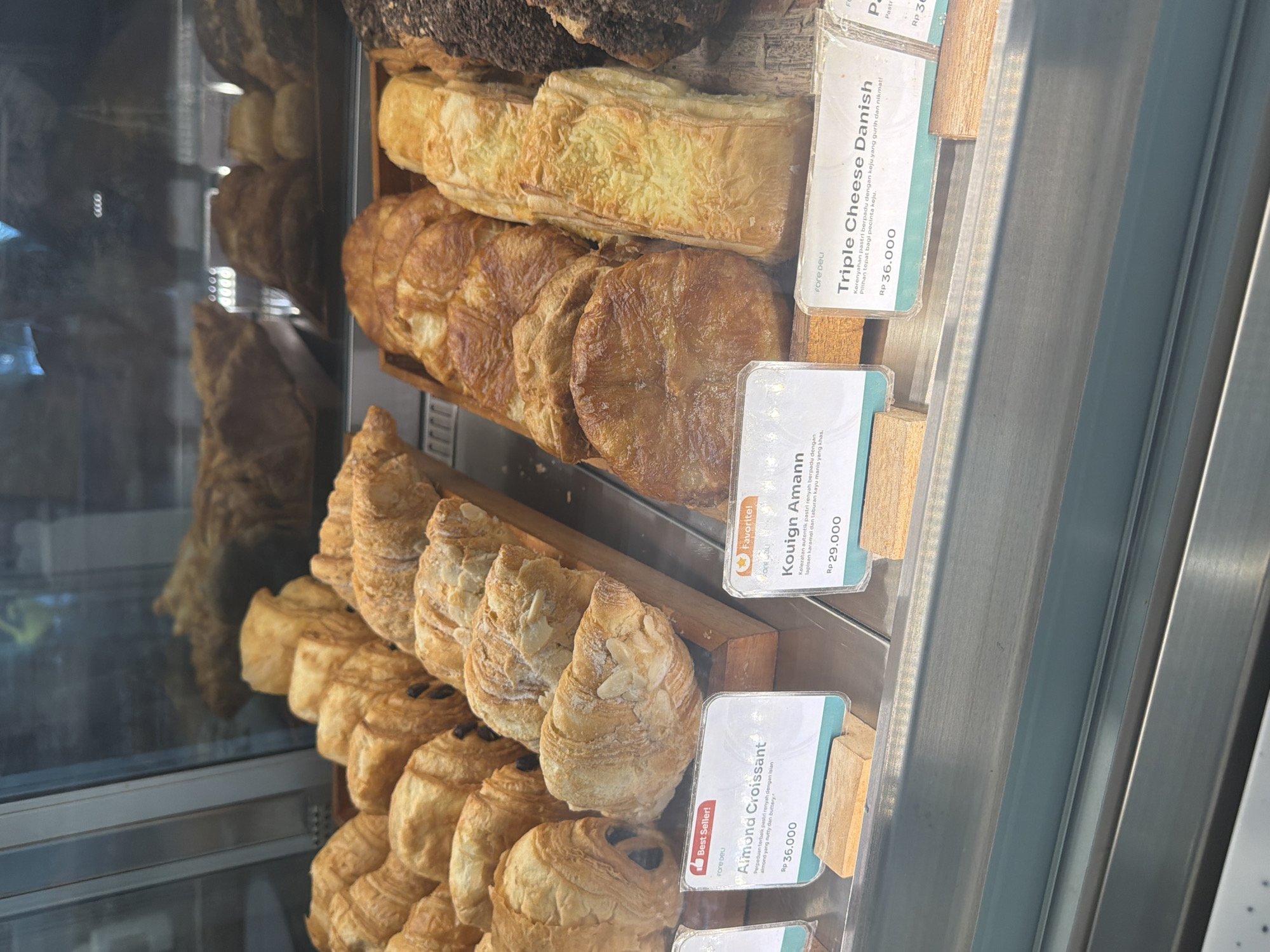 The image shows a close-up view of a display case filled with various pastries, likely in a bakery or cafe. The pastries are arranged on wooden shelves and in trays. Prominently displayed are croissants and danishes, some topped with almonds or cheese. Several price tags with product names are visible, including "Almond Croissant" priced at Rp 36,000, "Kouign Amann" at Rp 29,000, and "Triple Cheese Danish" at Rp 36,000. The display case has a reflective surface, showing some of the interior of the establishment and possibly other pastries in the background. The lighting is bright, illuminating the pastries. The scene is clean and organized, typical of a food service establishment. The image was likely taken in East Jakarta, Indonesia, given the currency (Rp for Indonesian Rupiah) and the likely context of such a bakery. No people are visible in the image.