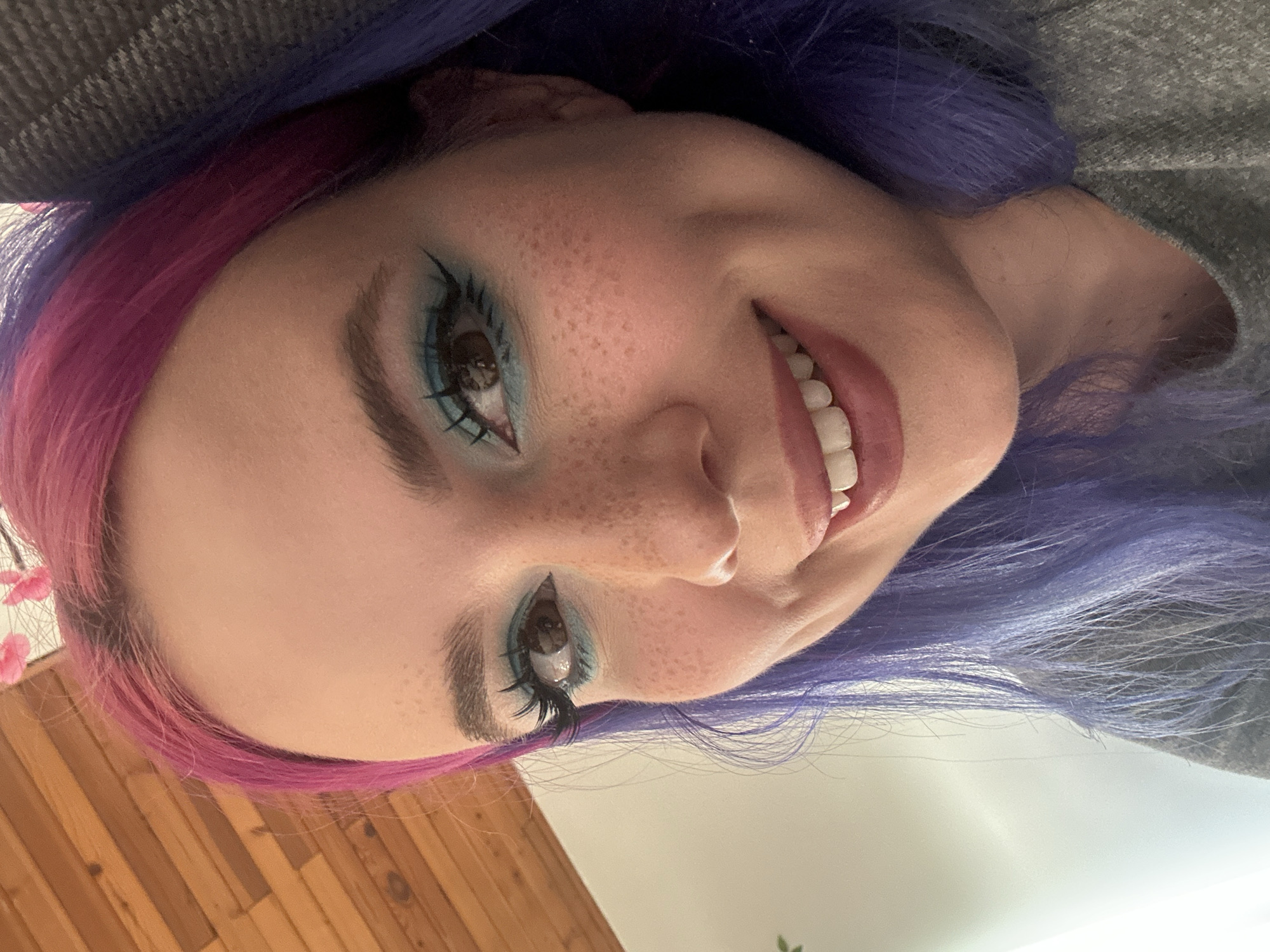 A close-up, angled shot of a woman with vibrant pink and purple hair, smiling. She has fair skin, freckles, and light brown eyes enhanced with blue eye makeup and false eyelashes. Her teeth are visible in a smile. She is wearing a gray top. A wooden floor is visible in the lower left corner. A light-colored wall and a hint of greenery are in the lower right corner. The location in the city of City of Zagreb, Croatia, is not determinable from the image.