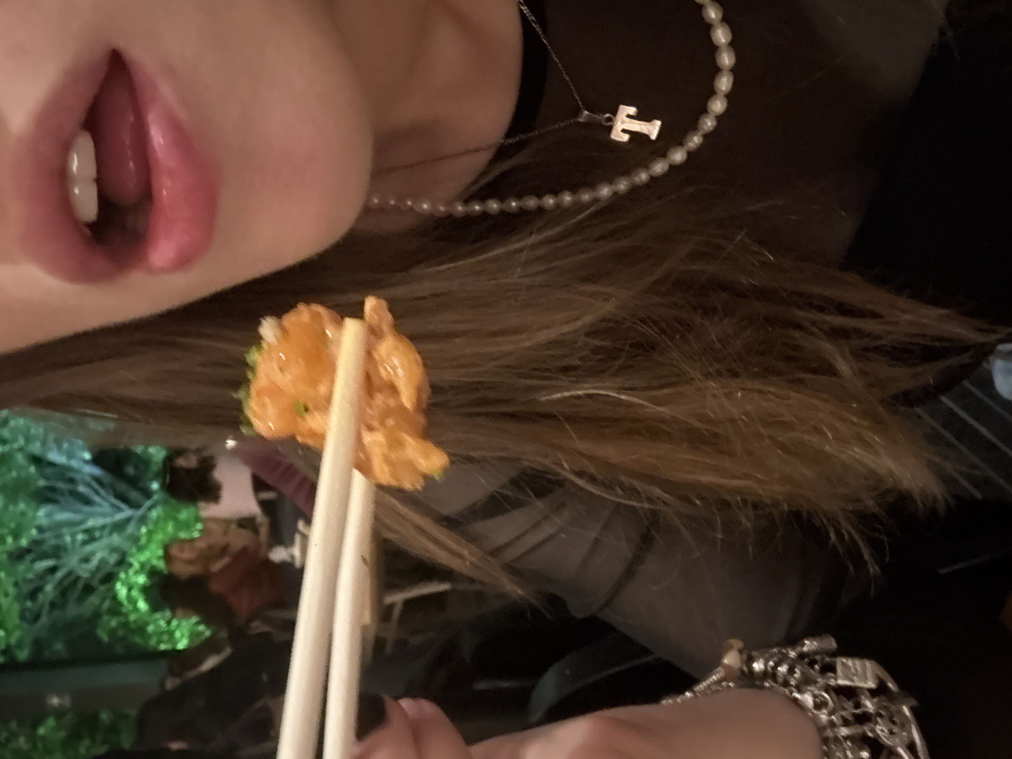 A person is holding food with chopsticks up to their mouth, presumably to eat. The person has a beaded necklace with a "T" pendant. The food appears to be some type of fried or coated seafood or meat. The person has brown hair and wears a metal bracelet. The surroundings show some vegetation and blurry shapes, suggesting an outdoor or restaurant environment, but the location cannot be specified from the picture.