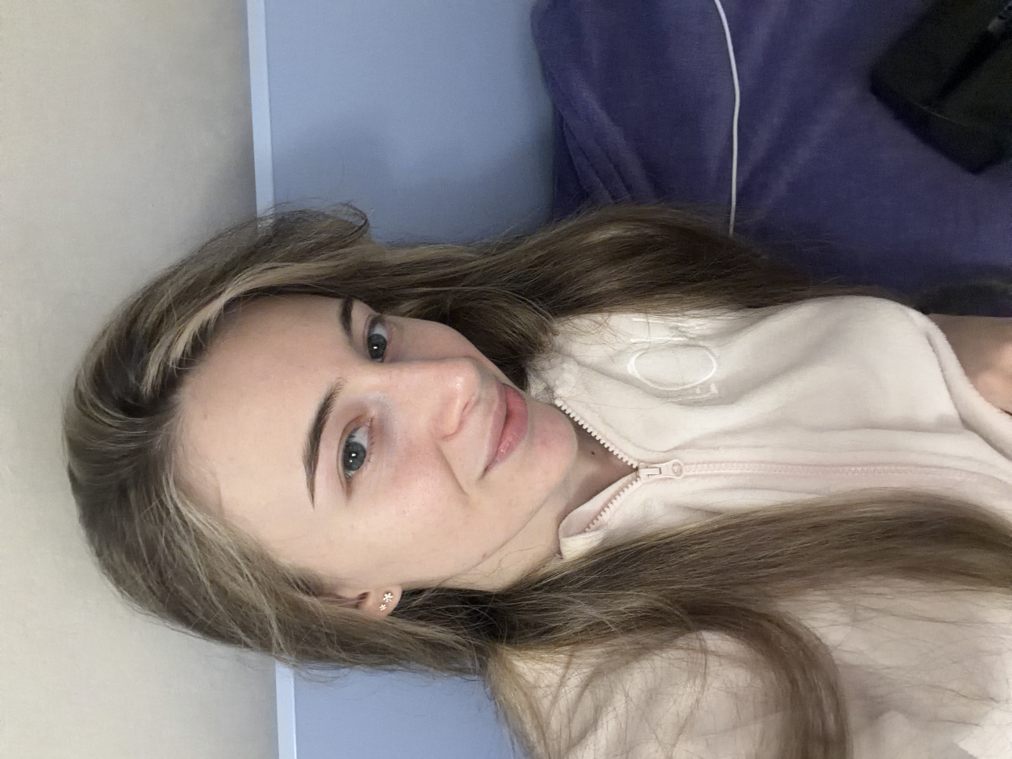 A young woman with light brown hair and blue eyes is lying down in a bed. She is wearing a light pink zip-up sweatshirt. A dark blue blanket is visible behind her. A black object is in the upper right corner of the image. There is no information available about the location in Plovdiv, Bulgaria.