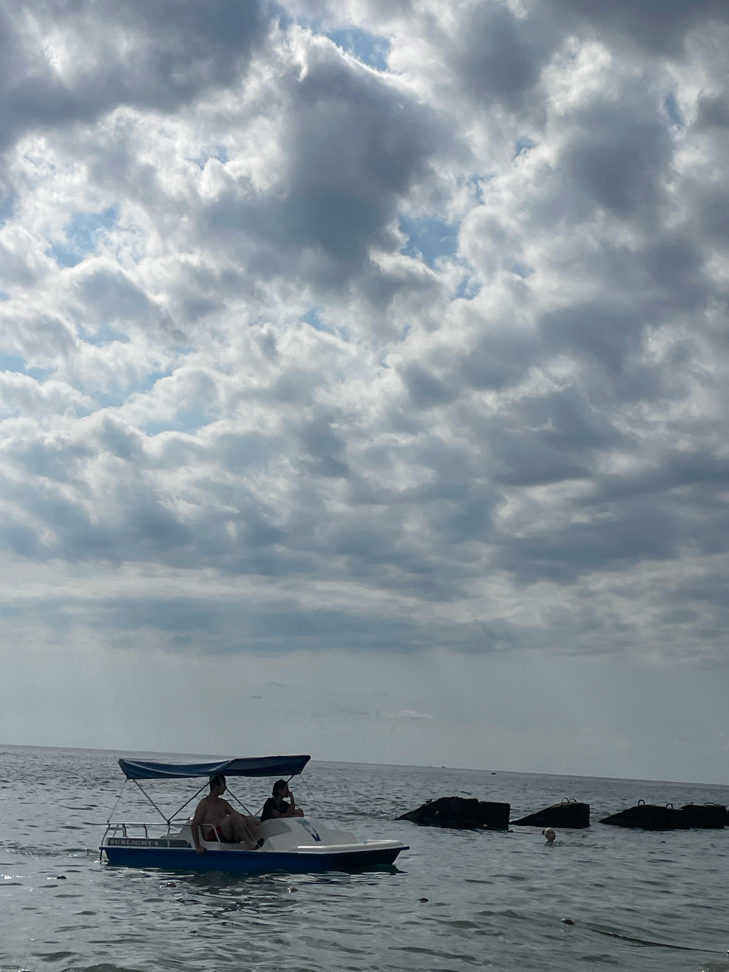 The image depicts a serene scene on the Black Sea coast of Sochi, Russia. In the foreground, a blue and white paddleboat, labeled "SUNLIGHT 4," glides across the water. Two individuals are seated in the paddleboat, one in the front and another behind. The sky above is a dramatic canvas of grey and white clouds, with patches of blue peeking through. In the distance, several dark, angular structures, possibly breakwaters or submerged objects, are visible, breaking the expanse of the sea. A lone person can be seen swimming in the water near these structures, adding a sense of human activity to the vast seascape. The overall atmosphere is tranquil, with the gentle ripples on the water and the vast cloudy sky creating a sense of calm and open space.