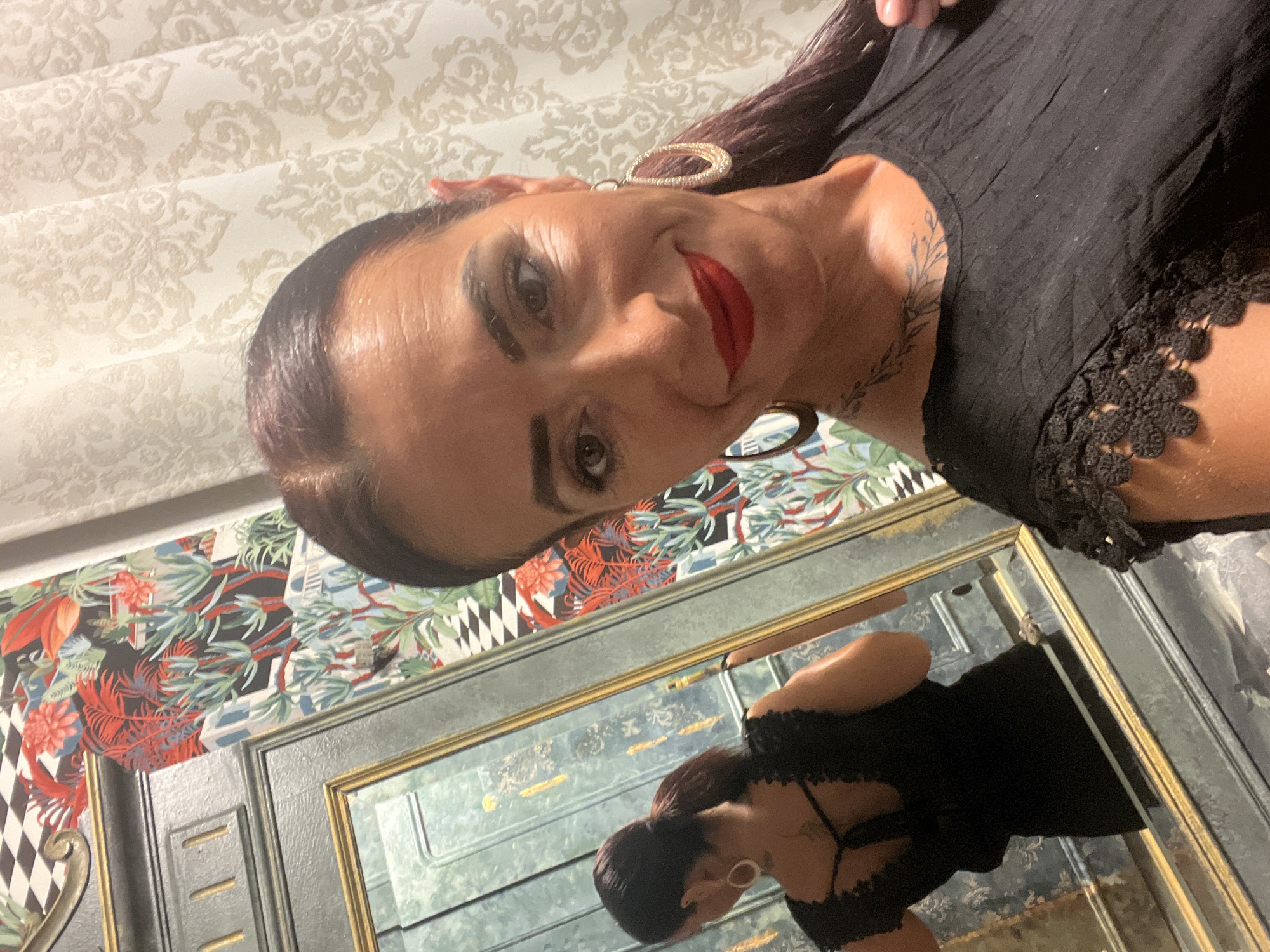 A woman with dark hair pulled back in a ponytail and wearing a black dress with lace trim takes a selfie in a room with ornate wallpaper and a large ornate mirror. The wallpaper features a bold floral and geometric pattern. The mirror reflects the woman's back, revealing a black dress with a lace-trimmed open back and a tattoo on her upper back. The woman is wearing large hoop earrings and red lipstick. The scene appears to be an interior setting, possibly a home or a hotel room, but no specific location in Konz, Germany can be identified from the image.