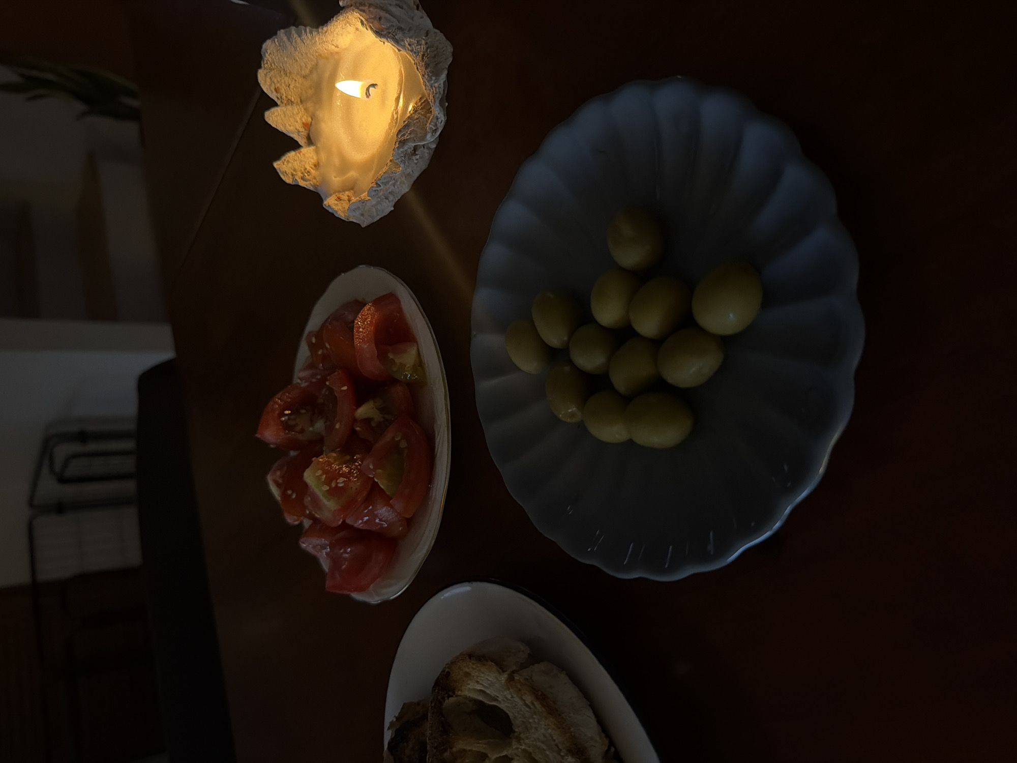 The image shows a table setting under dim lighting, likely illuminated by a candle. The table holds three dishes: one with sliced tomatoes, one with green olives, and one with pieces of bread. The tomatoes appear to be dressed with seeds. The objects are arranged on a dark brown surface. The specific location within Chang Phueak, Thailand is not discernible from the image. There are no people visible in the frame.