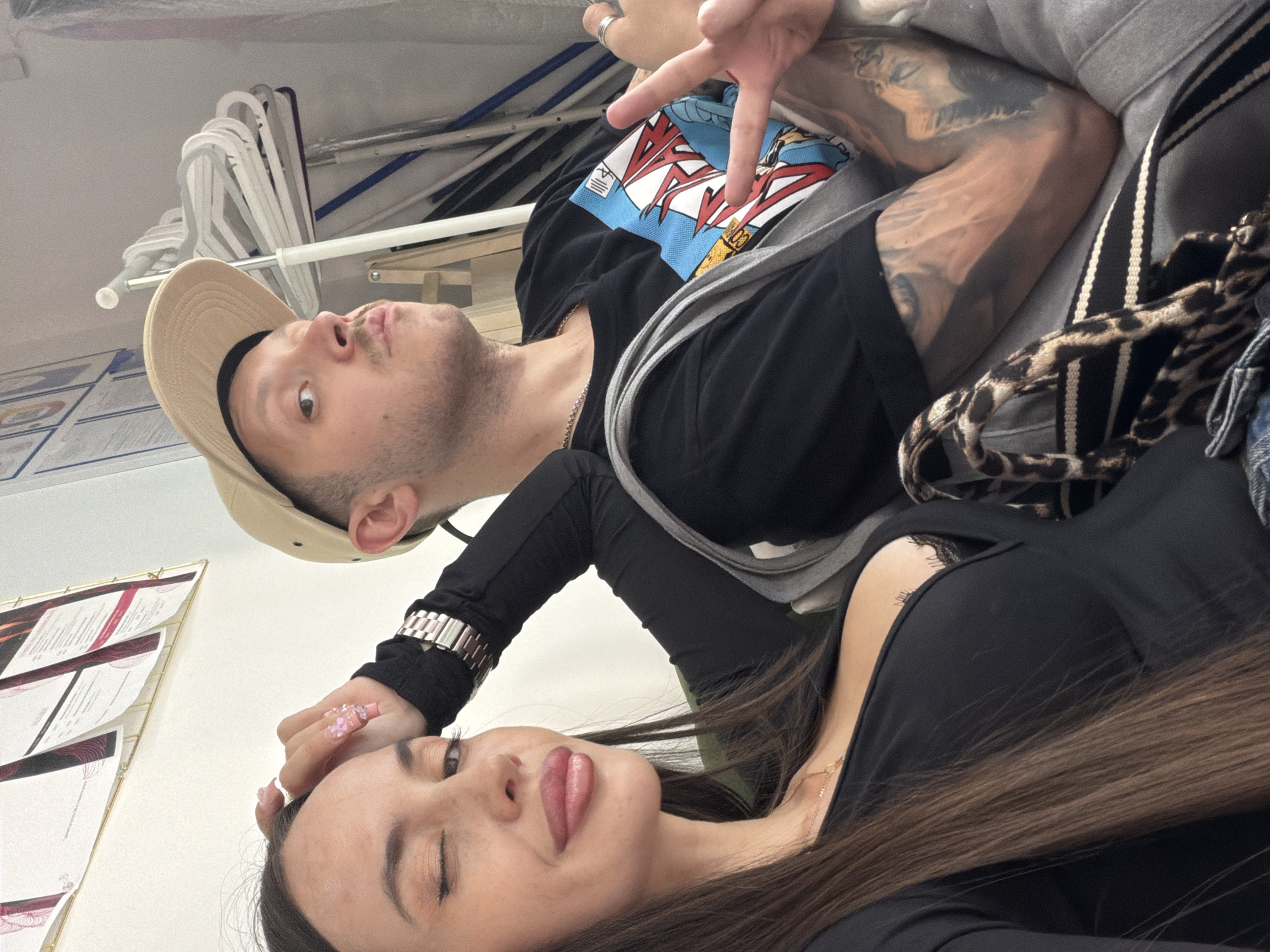 The image depicts two individuals, a woman and a man, posing for a selfie indoors. The woman, on the left, has long brown hair and is wearing a black top. She is winking and resting her head on her hand, which is adorned with decorative nails. A watch is visible on her wrist. The man, on the right, is wearing a beige baseball cap and a black t-shirt with a graphic print. He is puckering his lips and making a peace sign with his right hand. He has tattoos on his arm. Between them, a bag with a leopard print detail is visible. The background shows a white wall with framed documents and a rack of clothes hangers. The setting appears to be a room in Saint Petersburg, Russia, possibly an office or a dressing room, given the documents and hangers.