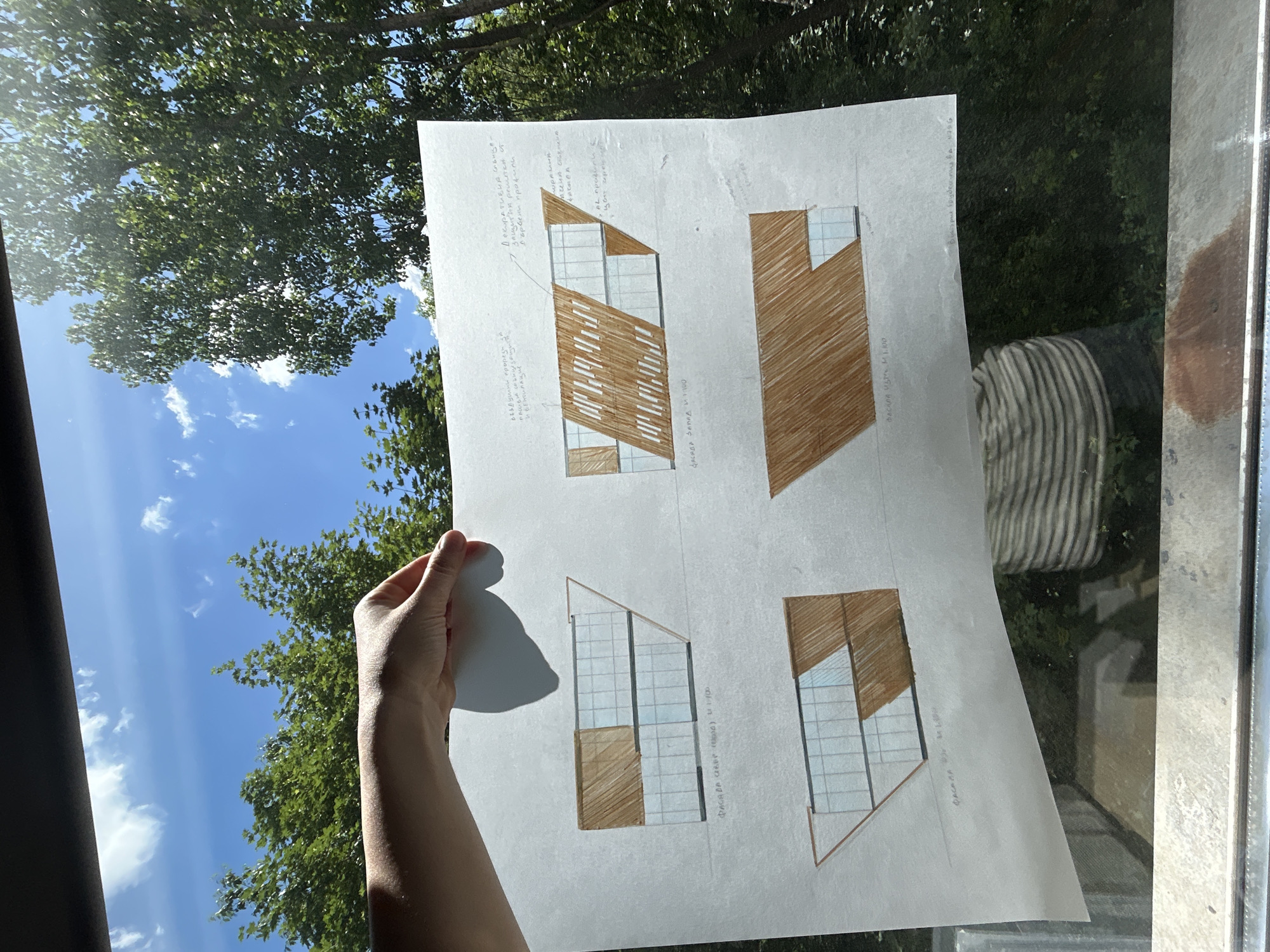 The image shows a hand holding a white sheet of paper with architectural sketches against a background of a tree canopy and a blue sky with clouds. The sketches depict building facades with a combination of grid-like window patterns and areas filled with brown shading, possibly indicating wooden elements. Written notes in a foreign language (possibly Bulgarian) are visible near each sketch, suggesting technical details or annotations about the design. The photo appears to be taken indoors, potentially through a window, given the reflection. The OCR mentions "Фасада запад М 1:100" which translates to "West facade M 1:100". The OCR also has Bulgarian words "Декоративна" which means decorative and "Вентилация" which means ventilation. The location is not depicted in the image, and Sofia, Bulgaria cannot be determined from the contents of the photo.
