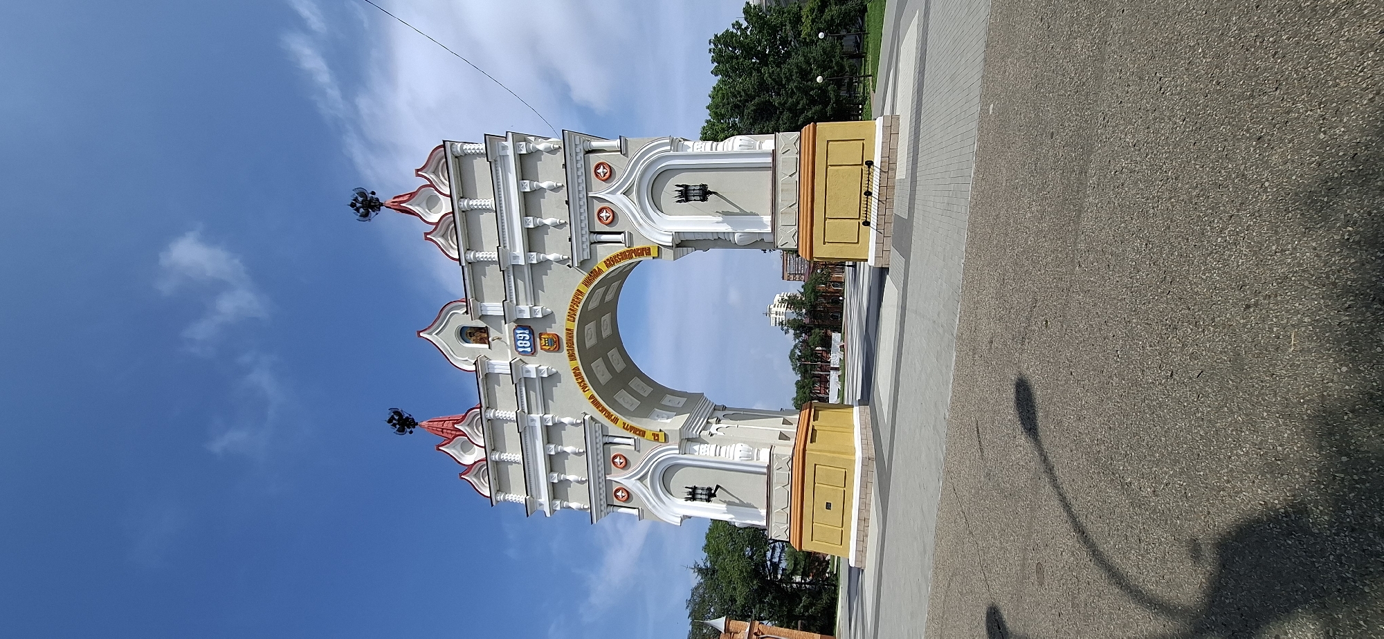 The image depicts the Triumphal Arch in Blagoveshchensk, Russia, a historical monument commemorating the 300th anniversary of the Romanov dynasty. The arch, constructed in gray and white with red accents, stands tall against a bright blue sky dotted with white clouds. It features ornate architectural details, including decorative columns, a central archway, and a crest with the year "1891" prominently displayed. Below the crest, a band of yellow lettering, likely an inscription, encircles the arch. The monument is situated in an open public space paved with interlocking gray and tan stones, suggesting a plaza or park area. In the foreground, a portion of a paved road with shadows from streetlights is visible. Trees line the perimeter of the plaza in the background, and several buildings, including a taller modern structure, are visible in the distance, indicating an urban setting. There are no people clearly visible in the frame.