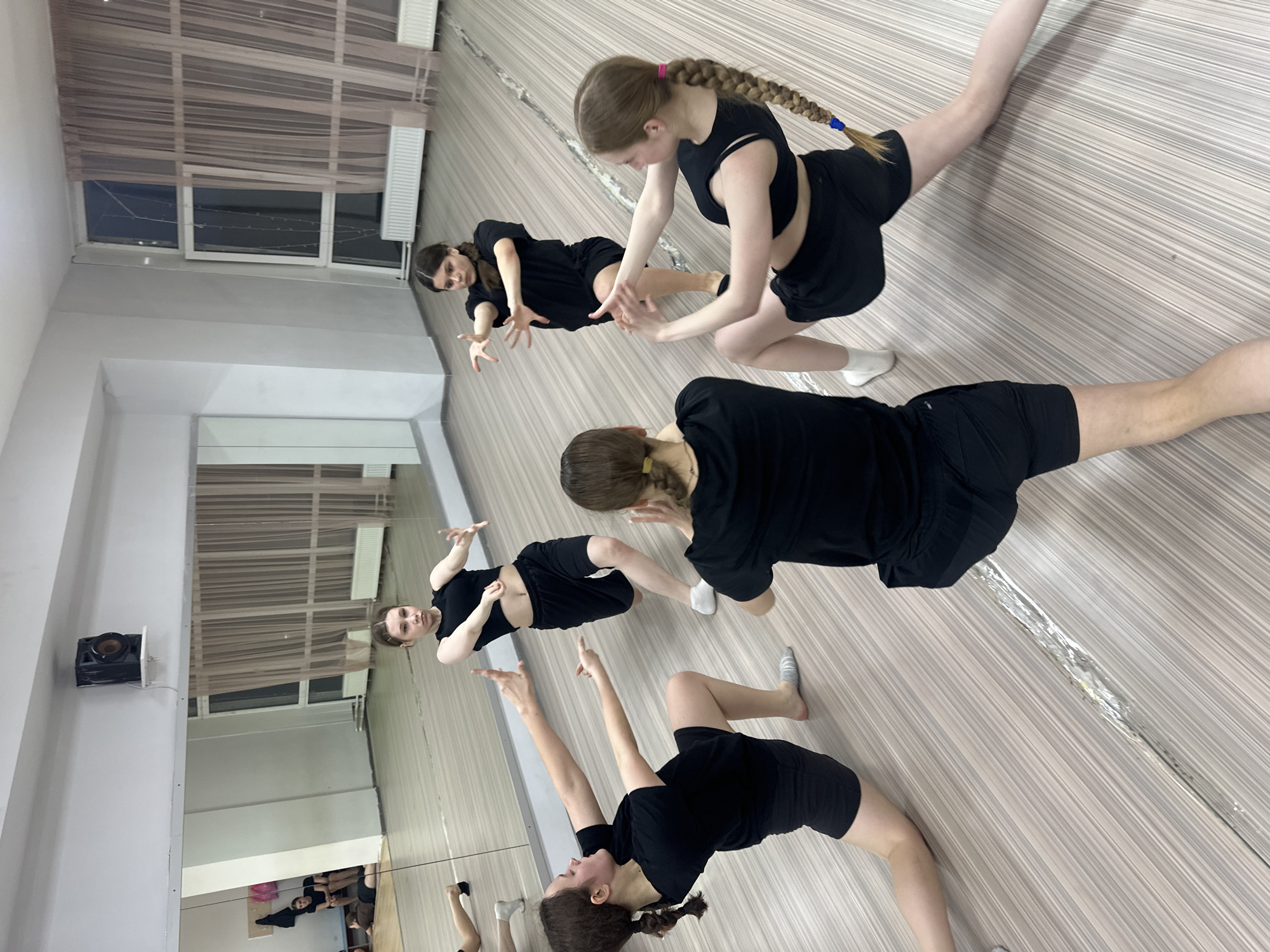 An indoor, tilted medium shot captures a group of young females engaged in a dance or exercise session within what appears to be a studio in Votkinsk, Russia. The room features a light wood-patterned floor and light-colored walls, with large mirrors covering one side, reflecting some of the participants.
There are at least five individuals visible directly or in reflection, all wearing matching black activewear consisting of shorts and short-sleeved tops or crop tops. Their hair styles vary, with some having braids (one with a blue tie, another with a pink tie) and others ponytails or buns. They are barefoot or wearing socks (some white, some striped grey). Their expressions are focused as they perform synchronized, stylized movements. One person in the foreground stretches with one leg extended and arms reaching back. Another in the middle ground is in a low, crouched position with hands extended. A third person is seen further back in a similar stance. The reflections in the mirror show other participants and further detail of the studio.
Notable details of the setting include large windows with light-colored sheer curtains or blinds, revealing natural light outside, suggesting it is daytime. Radiators are visible beneath the windows. A black speaker is mounted on a side wall. There is no discernible text in the image. The overall atmosphere conveys a practice or rehearsal session.