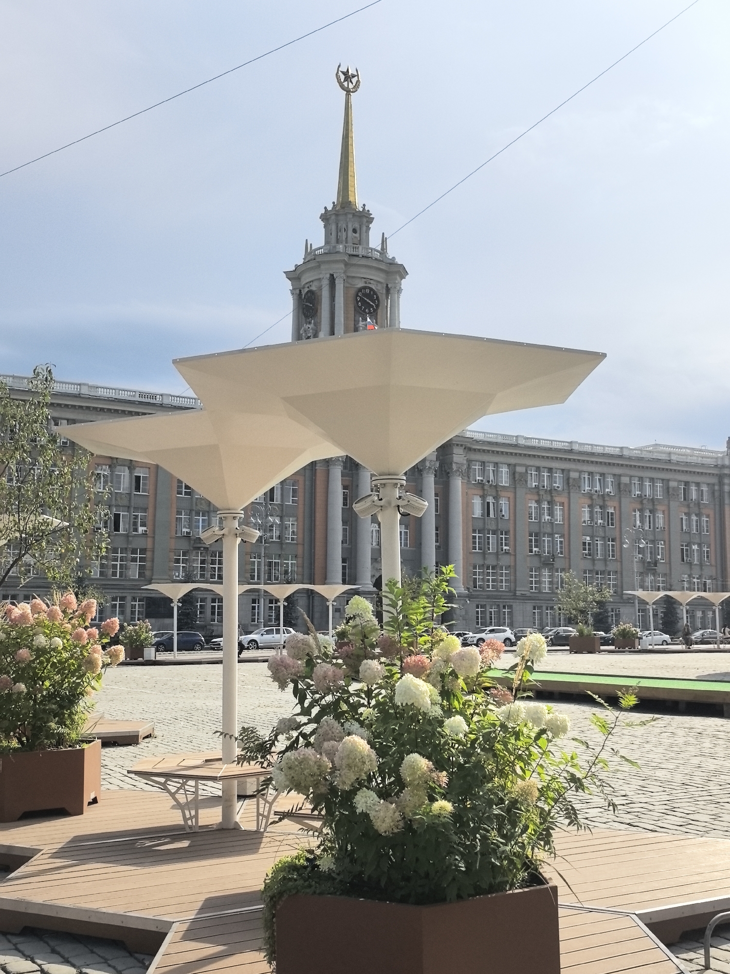 The image depicts the Square of the 1905 Revolution in Yekaterinburg, Russia, on a clear day. In the foreground, there are several large planters containing hydrangeas in bloom, with pink and white flowers dominating. These planters sit on a raised wooden platform, which also features a modern, geometric wooden table and bench. Behind the planters and wooden structures, large, modern white sunshades with angular canopies are visible.
Dominating the background is a grand, imposing building with a symmetrical facade, characterized by numerous windows and classical architectural elements such as columns. Atop this building, a prominent clock tower rises, culminating in a golden spire topped with a star and crescent symbol. The tower has large clock faces on at least two sides. The ground in front of the building and surrounding the square is paved with cobblestones. Several cars are parked on the street in front of the building, and a few figures are walking in the distance, too indistinct to identify. The sky is a pale blue with a few wispy clouds.