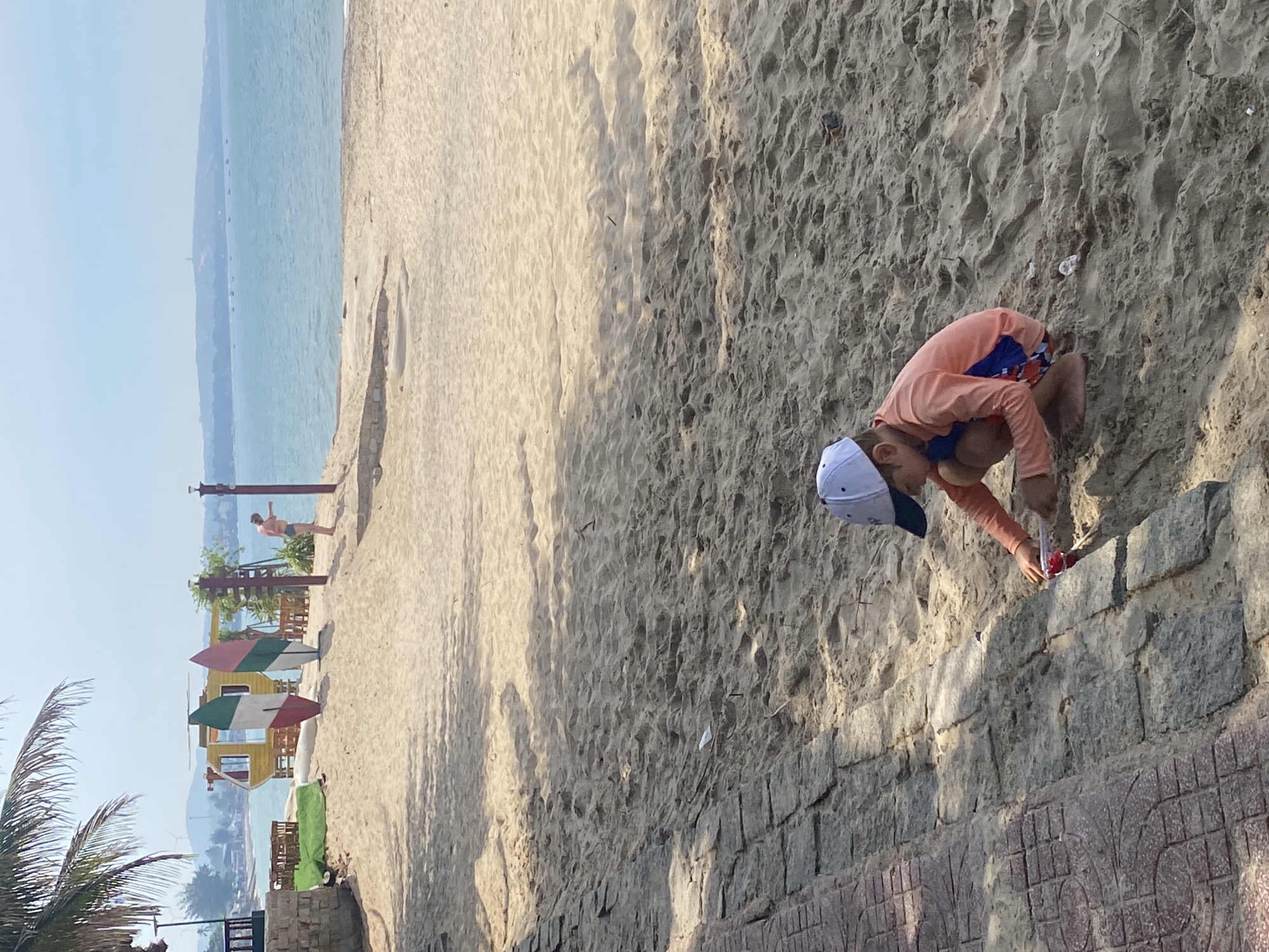 A child wearing a peach long-sleeved rash guard and blue and red patterned swim trunks is crouching in the sand on a beach. The child has on a white and navy blue baseball cap. In their right hand, the child is holding a small red object. The child is digging in the sand. In the background, the ocean is visible, with a distant shoreline and hazy mountains. There are several surfboards leaning against a colorful structure on the beach, as well as some wooden furniture. A palm tree overhangs the left side of the image. The sand has shadows cast on it from overhead objects, suggesting it is daytime. The scene is located on the beach in Phu Thuy Ward, Vietnam.