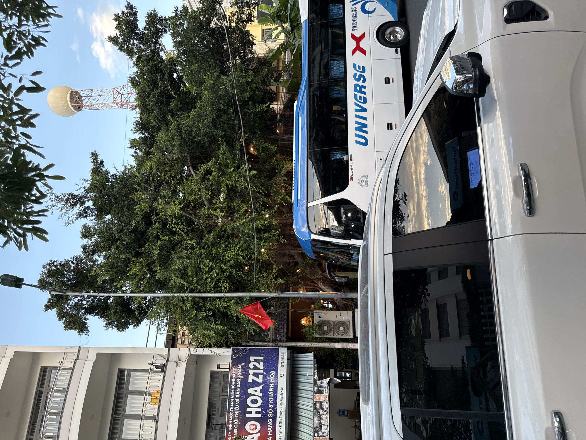 The image depicts a street scene in Nha Trang, Vietnam. In the foreground, the side of a white SUV is visible, with its polished side mirror reflecting the sky and trees. Behind the SUV, a white bus with blue accents and the word "UNIVERSE X" printed on its side is parked. A red Vietnamese flag is mounted on a pole near a large tree. To the left of the tree, a building with balconies and windows is partially visible. Further in the background, a tall, red and white tower with a large spherical object at its top stands against a clear blue sky. The scene appears to be set during the daytime, with natural light illuminating the surroundings.