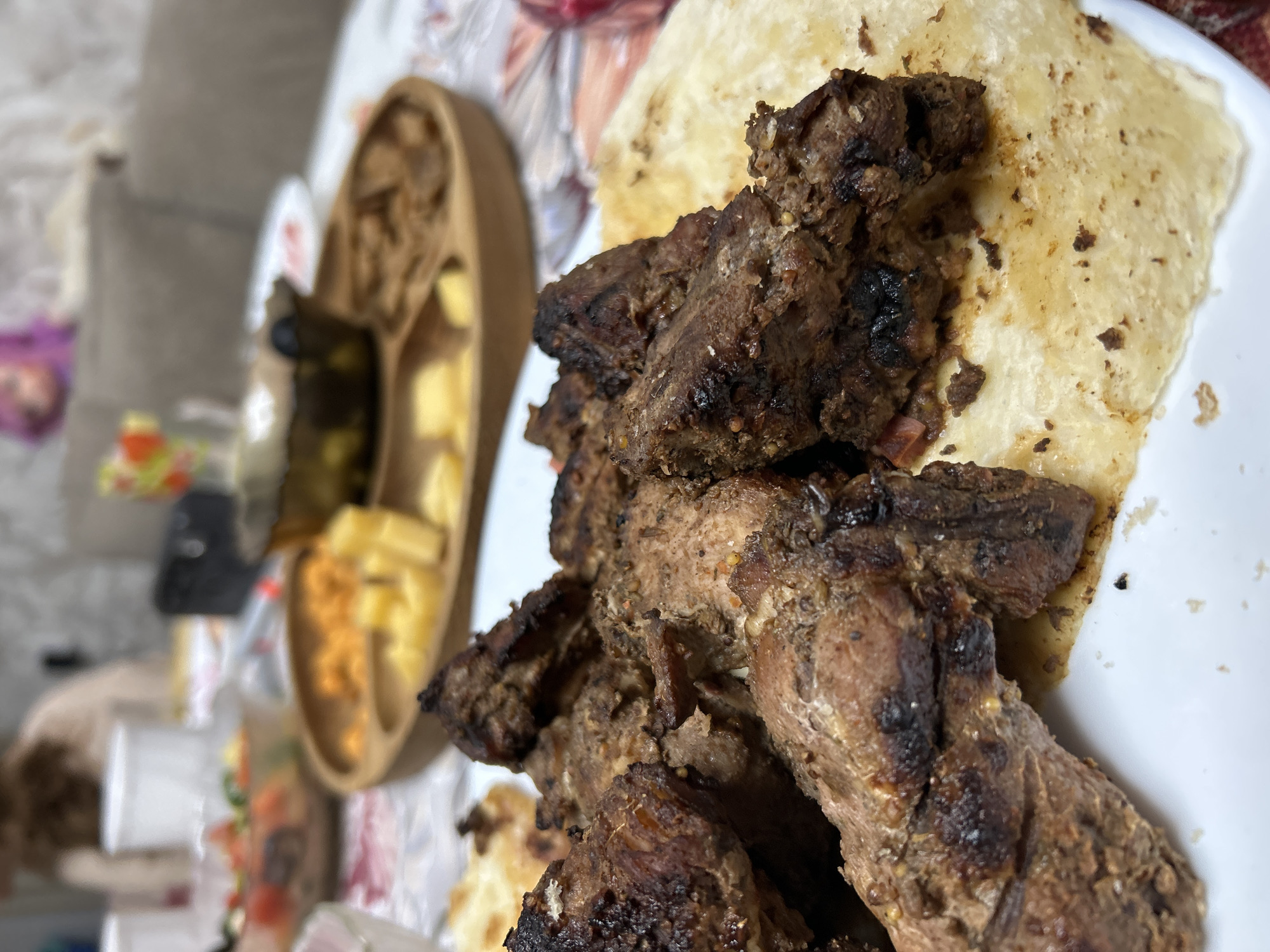 The image shows a close-up of food on a white plate. Specifically, there are pieces of grilled or roasted meat served with a flatbread or pita bread. In the background, slightly out of focus, is a wooden tray or serving dish with various snacks or appetizers like cheeses and olives. The surrounding environment appears to be a casual indoor setting. Noviy, Russia is not discernible from the image.