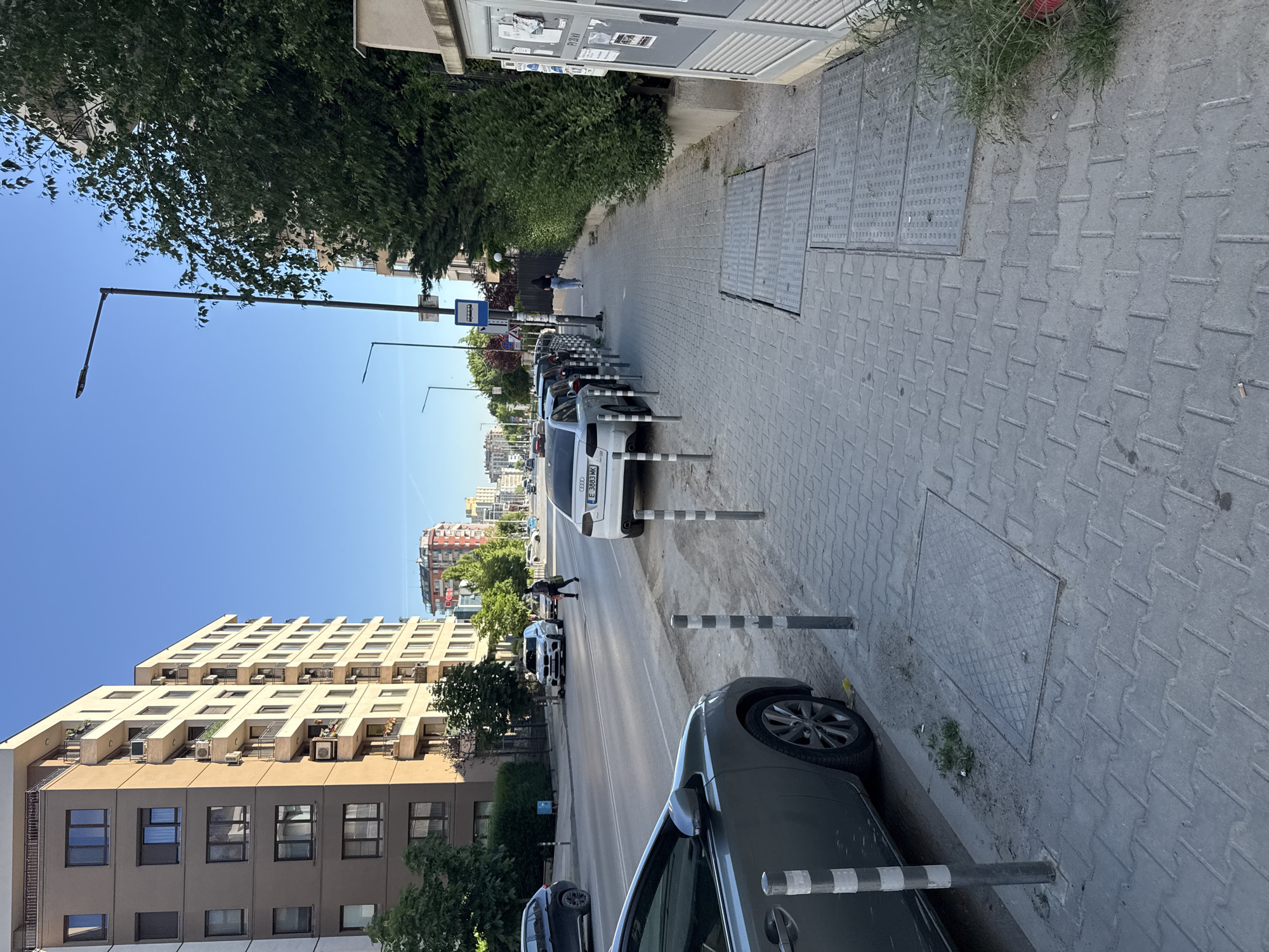 The image depicts a street scene in Sofia, Bulgaria. The view is of a road with parked cars along the right side, separated from the sidewalk by striped metal bollards. A person is walking across the road, towards the parked cars. The architecture includes a large, multi-story building on the left side of the street with repeating patterns of windows and balconies, as well as other buildings in the distance. A street lamp is visible on the left side of the street. The sidewalk is paved with gray interlocking blocks. The sky is clear and blue.