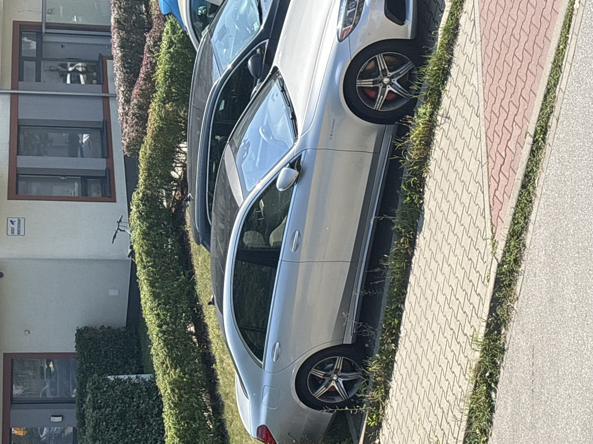 The image depicts a silver Mercedes-Benz S-Class sedan parked on a paved surface adjacent to a building and a manicured hedge line. The car is positioned on the right side of the frame, facing towards the right. A black car is partially visible behind it, and a portion of another vehicle is visible in the upper right corner. The building has a light-colored facade with windows and a sign that reads "OBIEKT MONITOROWANY" (Object Monitored), suggesting a security presence. A bicycle is propped against the building in the background. The ground is paved with interlocking bricks, with grass growing in the cracks and along the edges. The scene appears to be an outdoor parking area in a residential or commercial district. There are no individuals visible in the image. The presence of the sign and the parked vehicles indicates a developed urban setting.