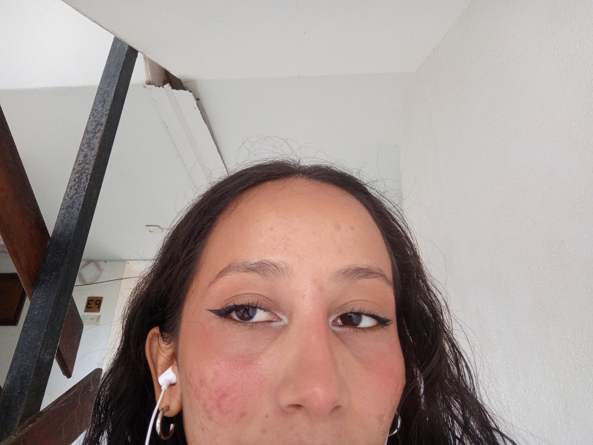 The image shows a close-up of a person's face from a low angle, possibly taken from below. The person has brown hair and brown eyes. They are wearing eyeliner and possibly blush. A white earbud is visible in their left ear along with a silver hoop earring. Their skin appears to have some redness. In the background, there is a white wall and a section of stairs with a dark metal railing. Based on the presence of Spanish writing on the stairway wall, it is plausible that the photograph was taken in the city of Porlamar, Venezuela.