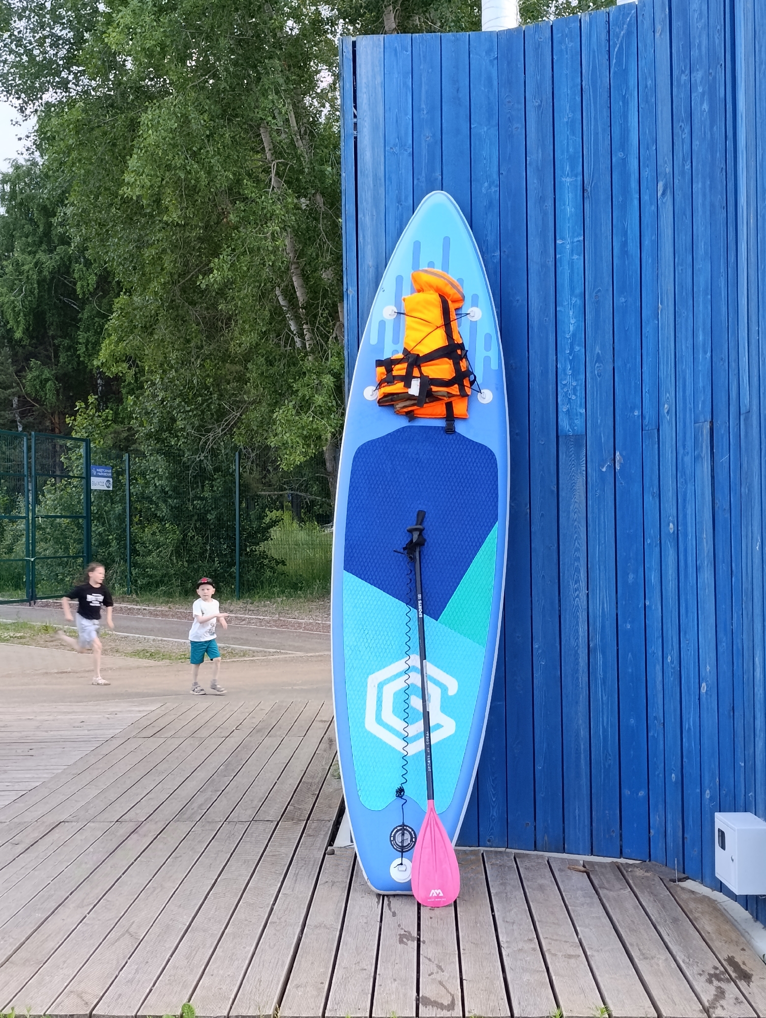 An upright blue paddleboard leans against a bright blue wooden fence. The paddleboard is decorated with a blue and light blue geometric pattern and has an orange life vest strapped to its surface. A pink paddle rests against the bottom of the paddleboard. Two young boys are in the background, on a paved path, with a green fenced area behind them. One boy is running and the other is standing. The scene appears to be outdoors, possibly near a body of water, given the paddleboard. The trees in the background suggest a natural setting. The time of day appears to be daytime, possibly late afternoon or early evening, given the lighting. A white electrical box is mounted on the blue fence near the bottom right of the image. The text "SPORTS IN ORBIT" is visible on the paddle shaft.