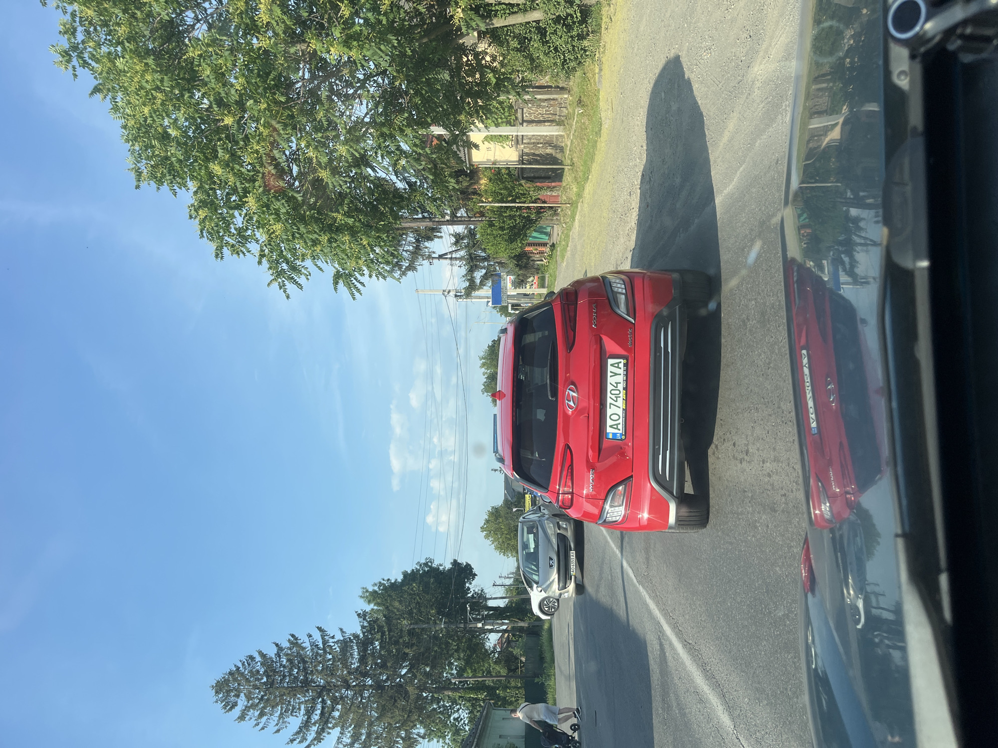 This is a street-level, rear-view shot taken in Mynai, Ukraine. The focal point is a red Hyundai Kona electric vehicle in the foreground, viewed from behind. The vehicle has a license plate that reads "AO 7404 YA". To the left of the Hyundai, a silver Peugeot is visible, its license plate reading "AO 6353 AX". Further back on the left, a white sedan is parked. On the sidewalk to the left, a person with a stroller is walking away from the camera. The scene is a paved road with a gravel shoulder. Houses with fences line the right side of the road, and trees, including a tall conifer and leafy deciduous trees, are present. The sky is clear blue with scattered white clouds. The shot is taken from inside another vehicle, with the hood and windshield of the camera car visible in the bottom of the frame.